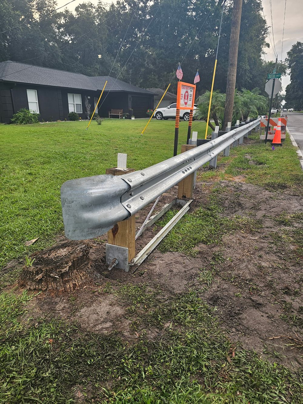 Damaged roadside guardrail with bent end section and work cones along a wet shoulder.