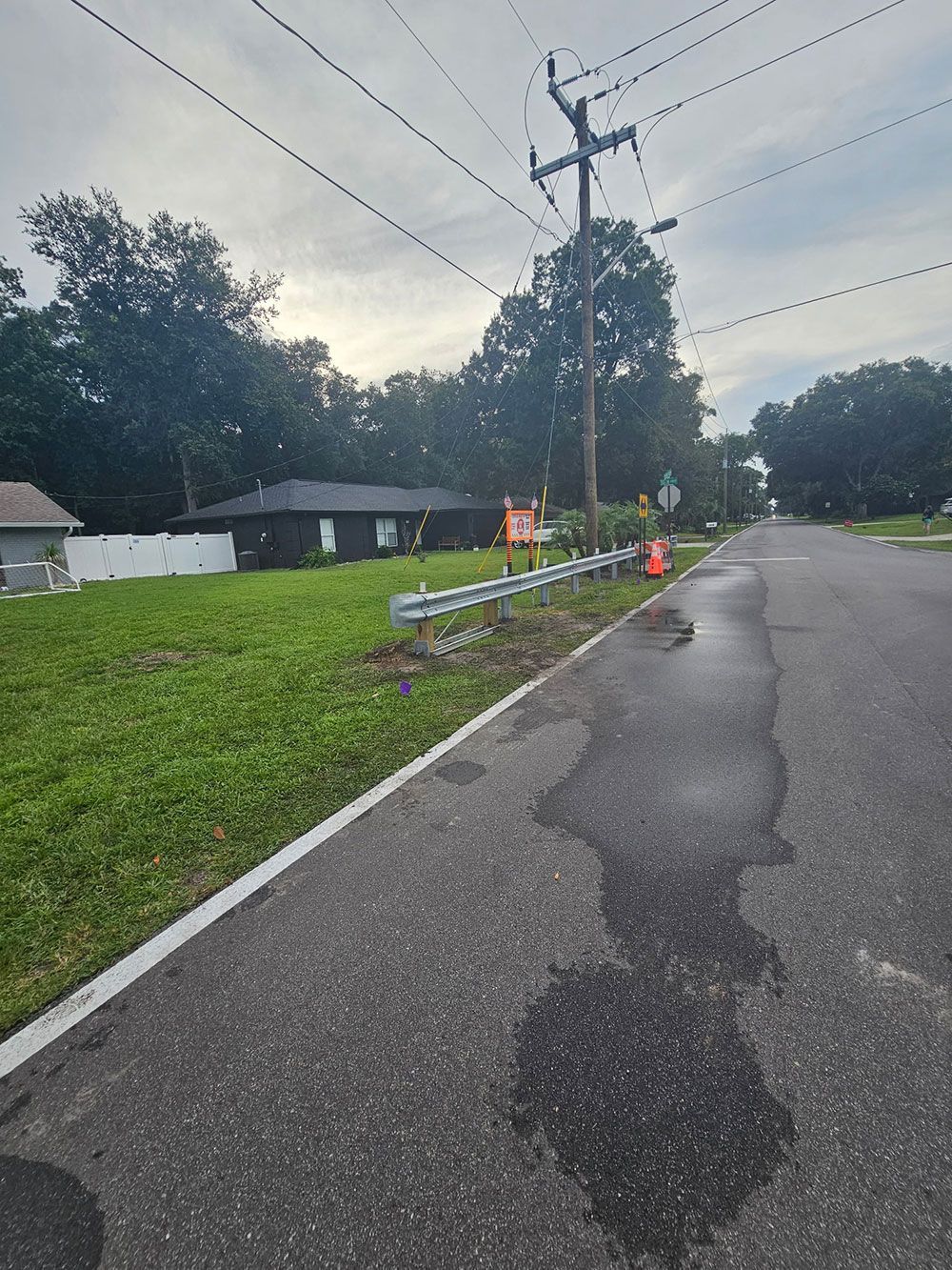 Damaged roadside utility pole with cones and caution tape along a wet residential street at dusk.