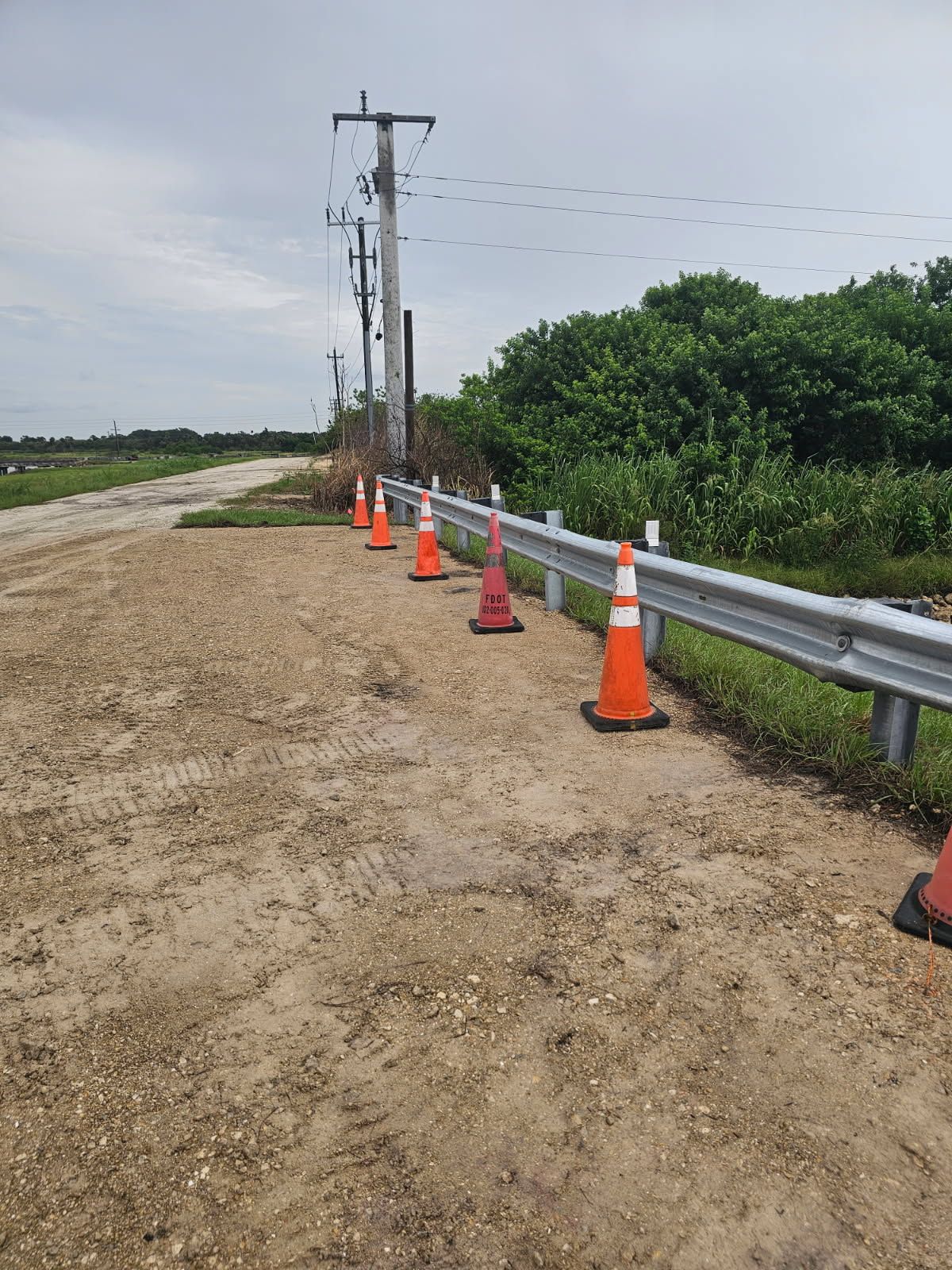 Rural dirt road beside a metal guardrail with orange traffic cones and utility poles along the roadside