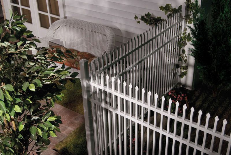 White picket fence beside a house, with plants and a covered bench in a small garden at night