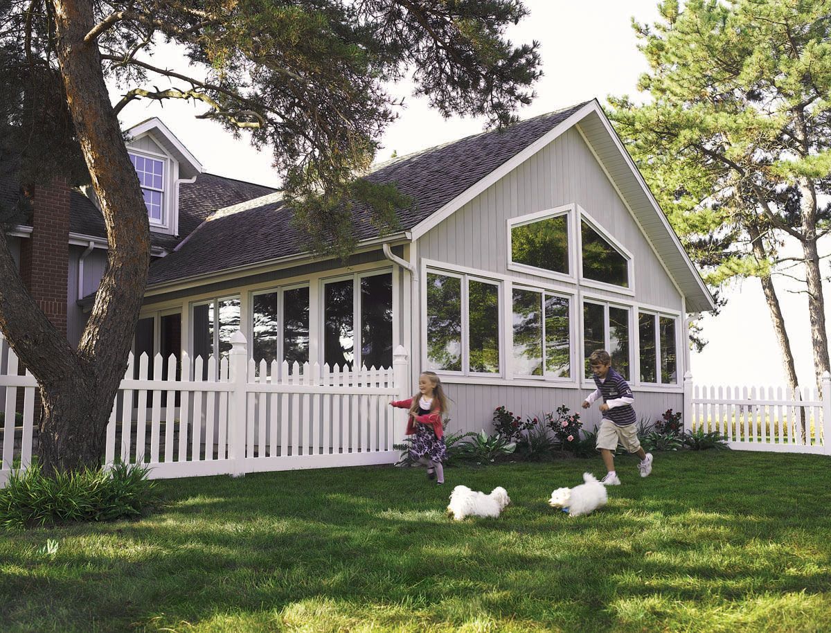 White house with large windows, children on a porch, and two white ducks on a grassy lawn.