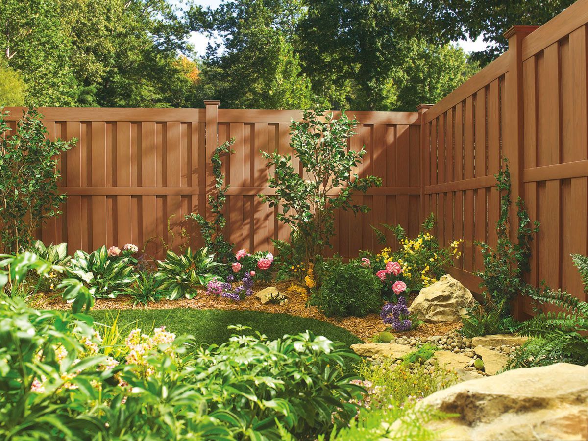 Wooden backyard fence with lush green plants, flowers, and stone landscaping in a sunny garden.