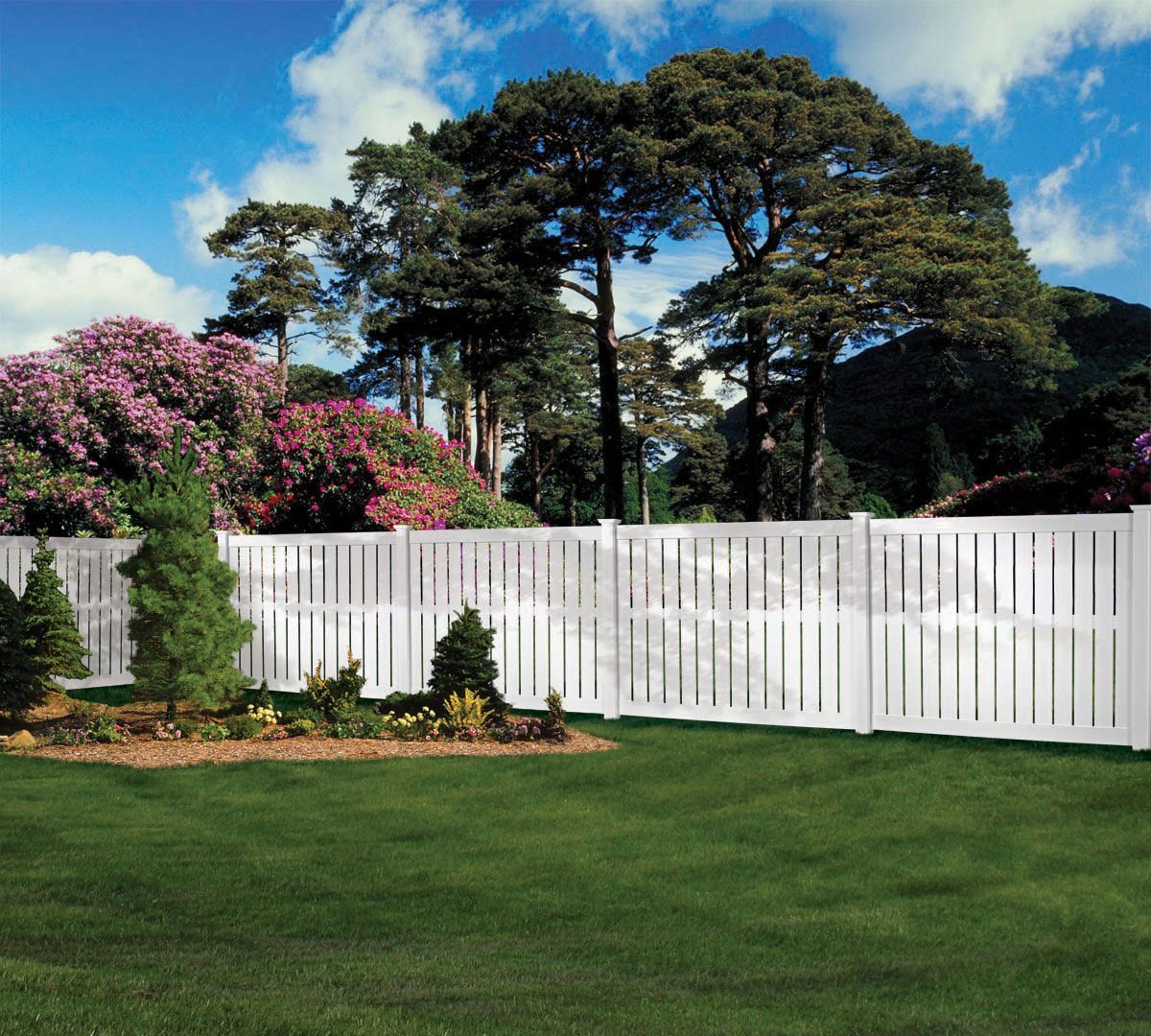White vinyl fence along a green lawn with shrubs and tall trees under a blue sky