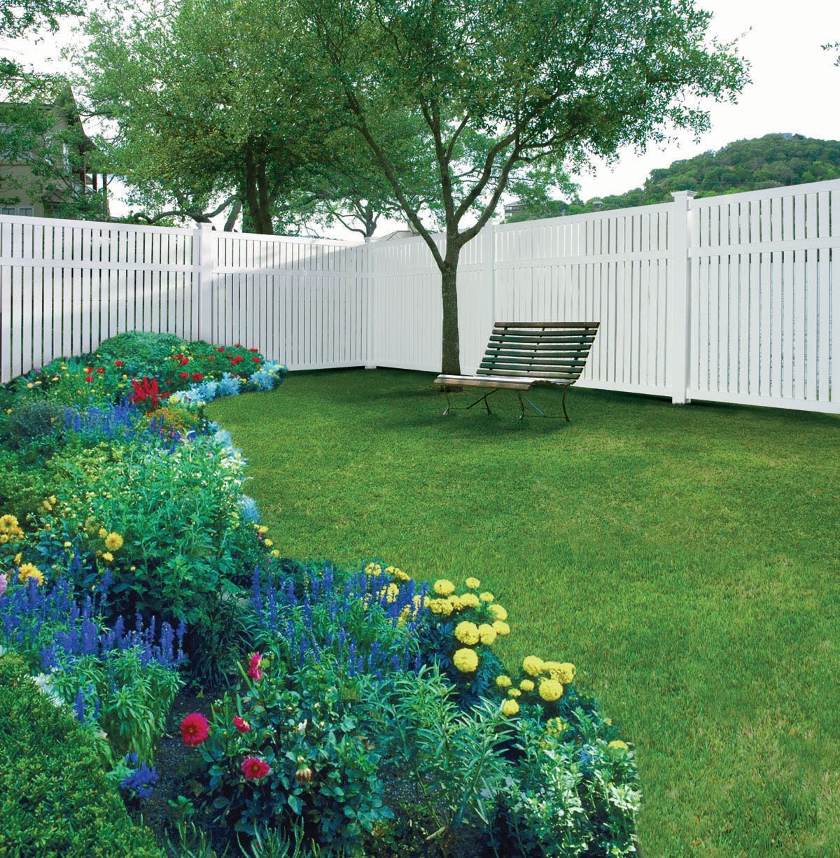 Flower-lined backyard with green lawn, white fence, and a tree with a bench underneath.
