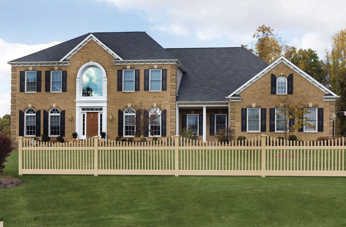 Large brick suburban house with black roof, white fence, and a green lawn under a blue sky