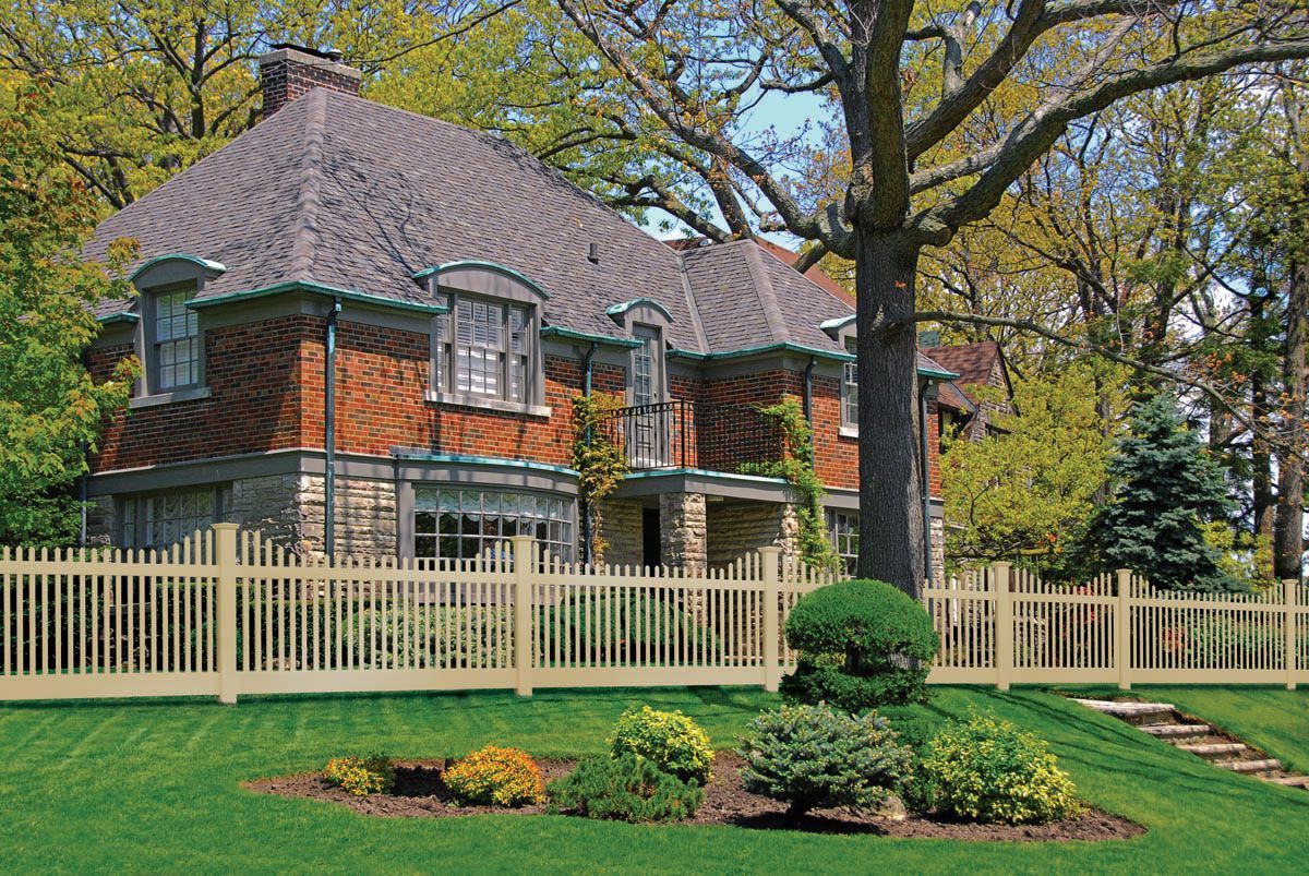 Brick house with a gray roof behind a cream fence, surrounded by green lawn and trees.