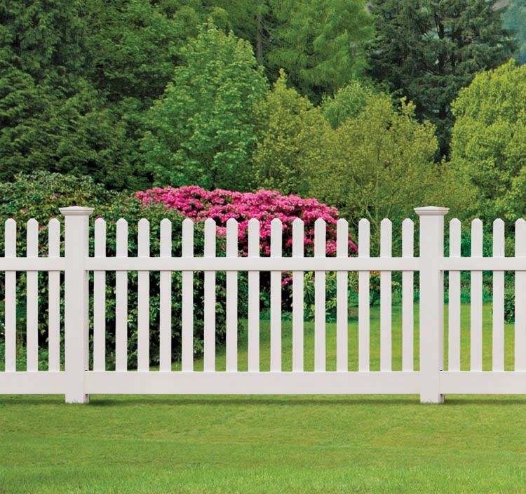 White picket fence in a green yard with pink flowers and dense trees behind it