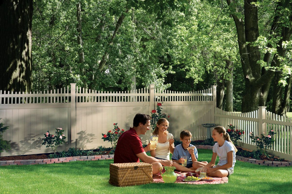 Family sitting on a picnic blanket in a sunny park with a white fence and trees behind them