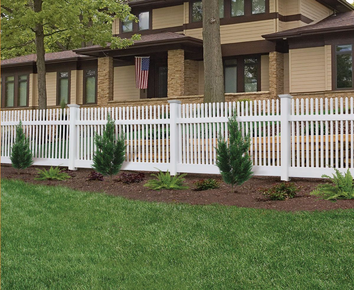 White vinyl fence in front of a tan house with shrubs and green lawn.