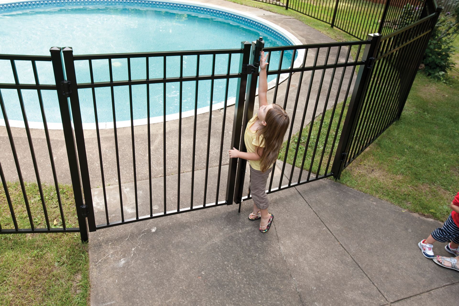 Child standing at a black fence beside a swimming pool, reaching toward the gate