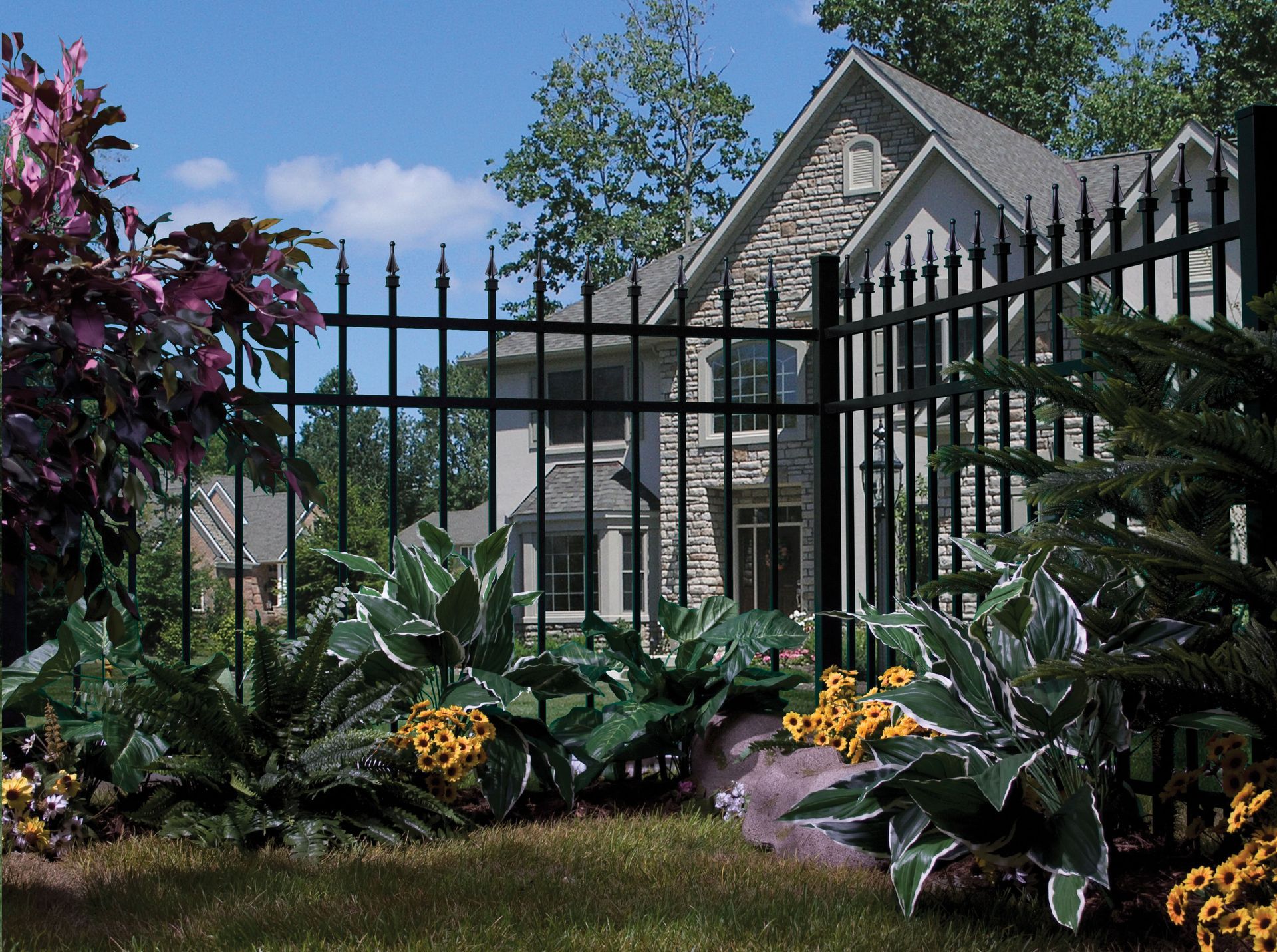 Black iron fence in front of a stone house, with green shrubs and yellow flowers under a blue sky.