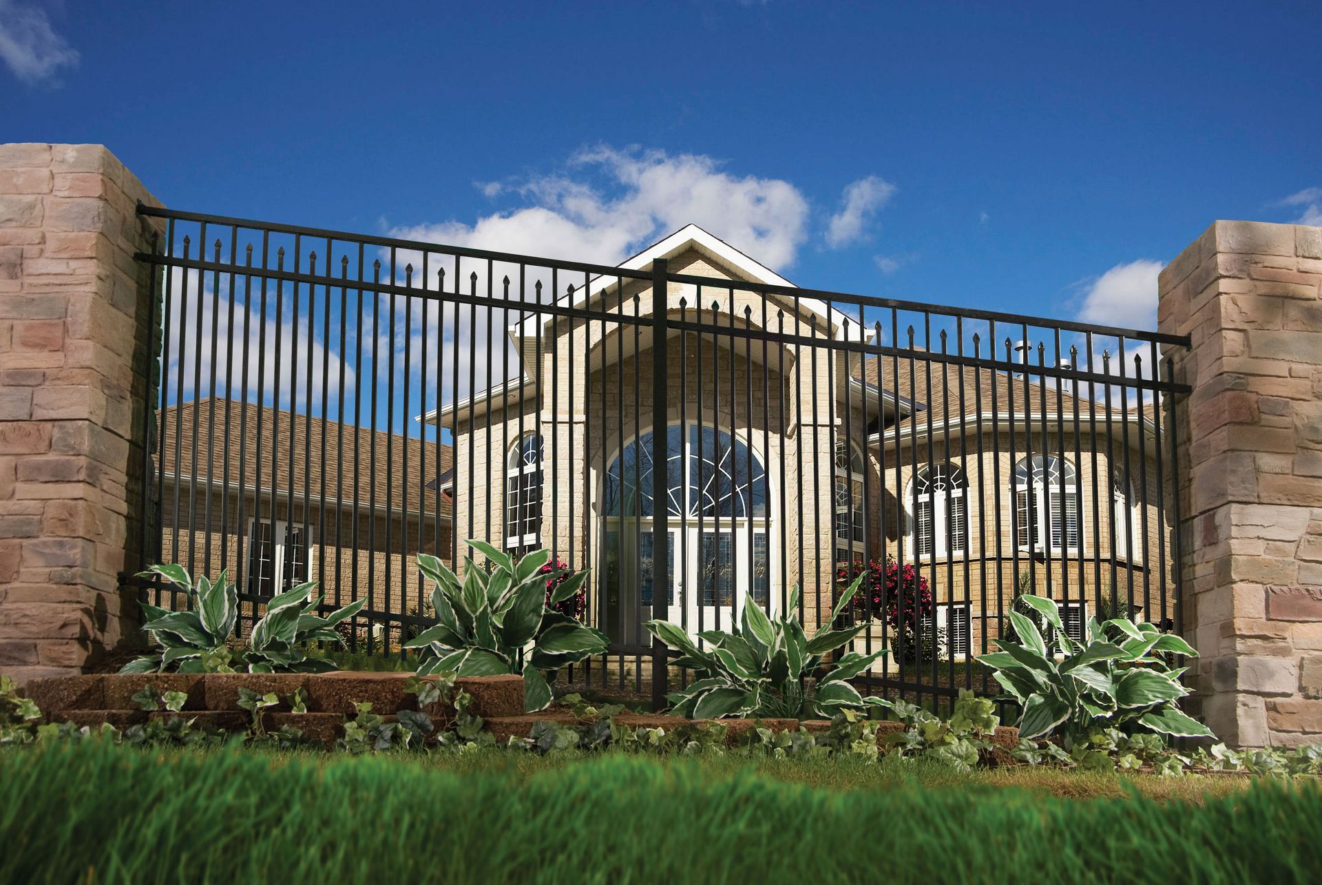 Black metal gate in front of a beige house with stone pillars, landscaped garden, and blue sky