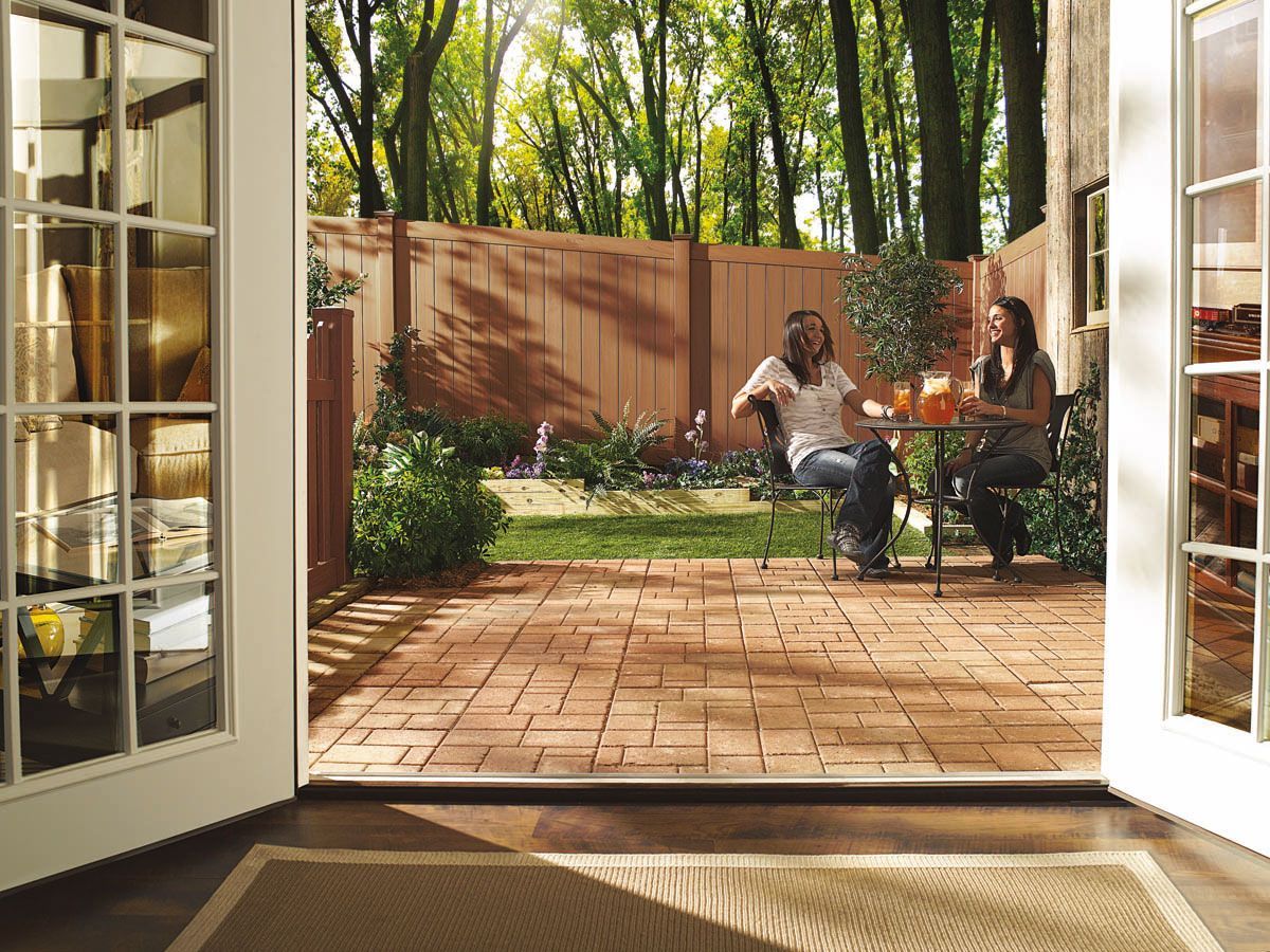 Open double doors to a brick patio with two people seated at a table in a sunlit garden.