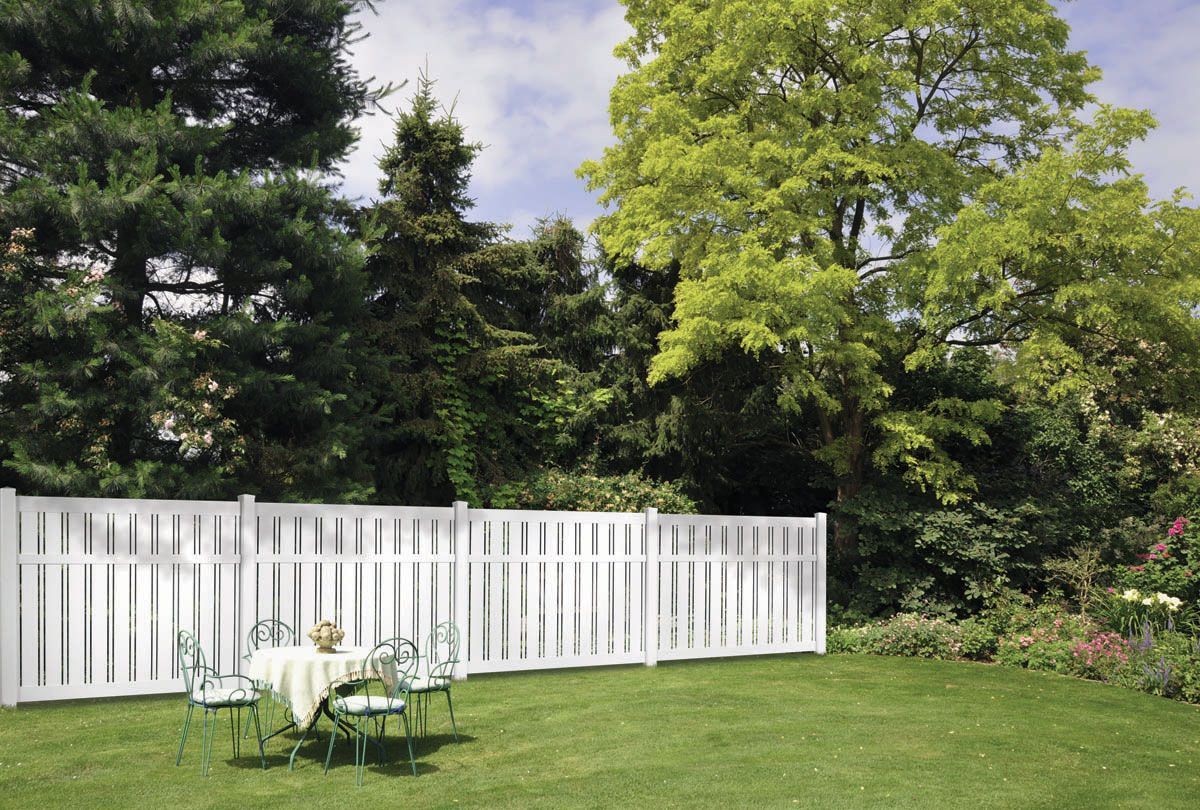 White garden fence beside a green lawn and trees with a small table and chairs in the foreground