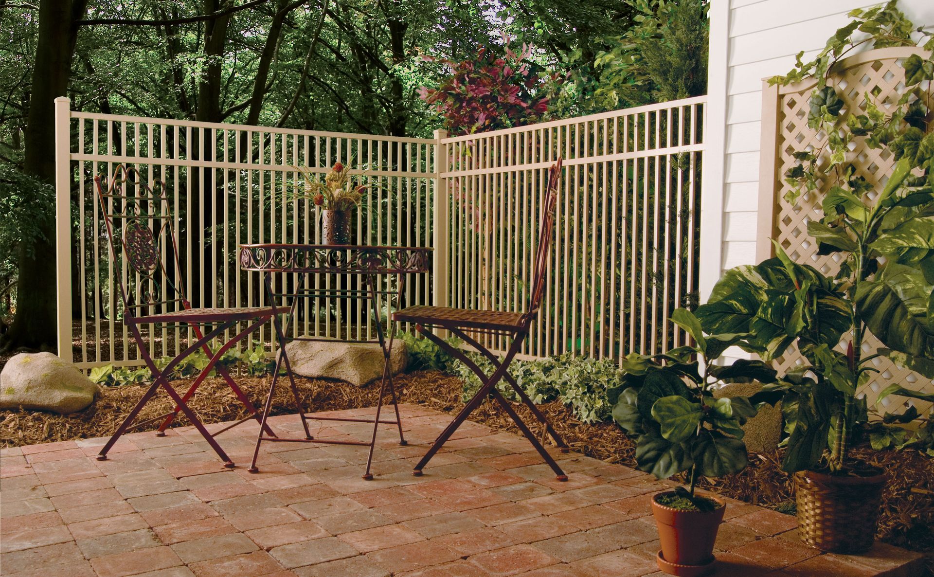 Patio garden with two folding chairs and table, brick floor, potted plants, and a white fence backdrop
