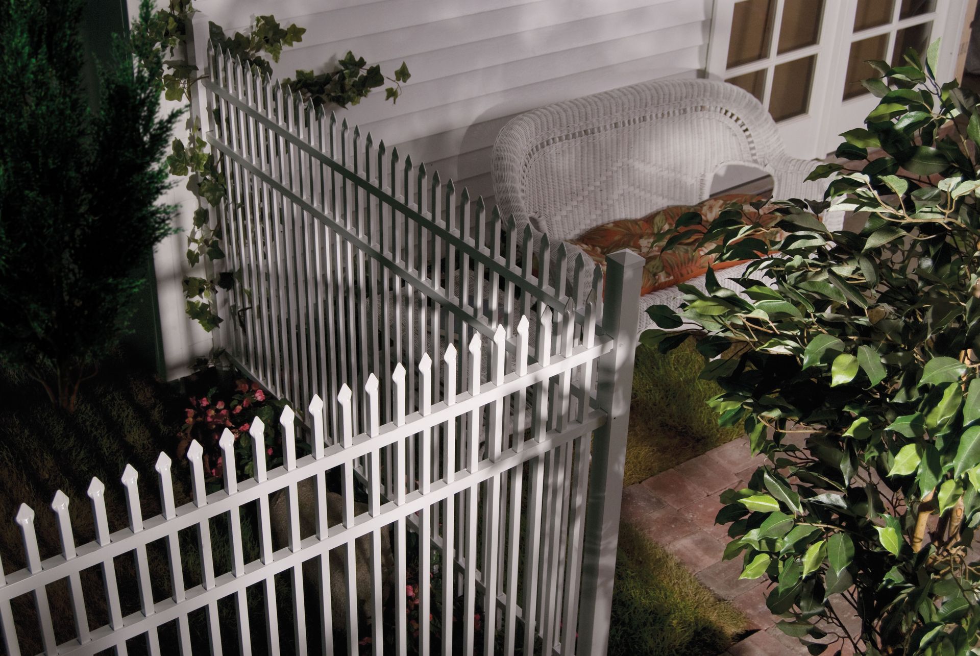 White metal fence by a house with a lit walkway and dense bushes at night
