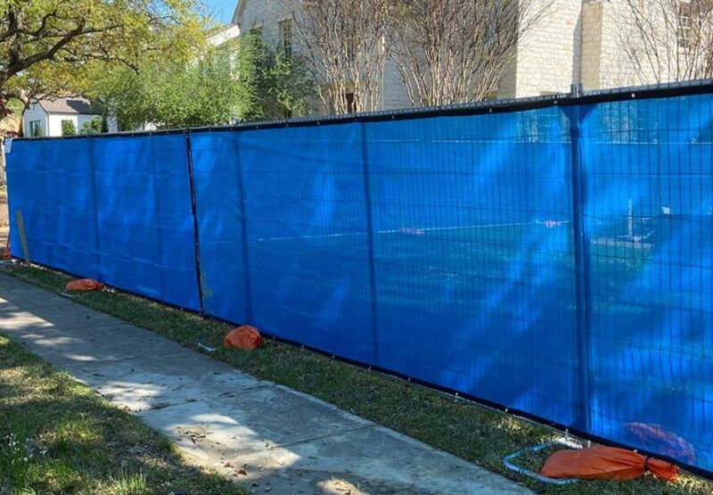 Blue construction fence along a sidewalk with trees and buildings in the background