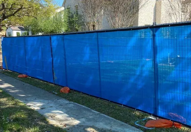 Blue construction fence along a sidewalk beside a building and trees