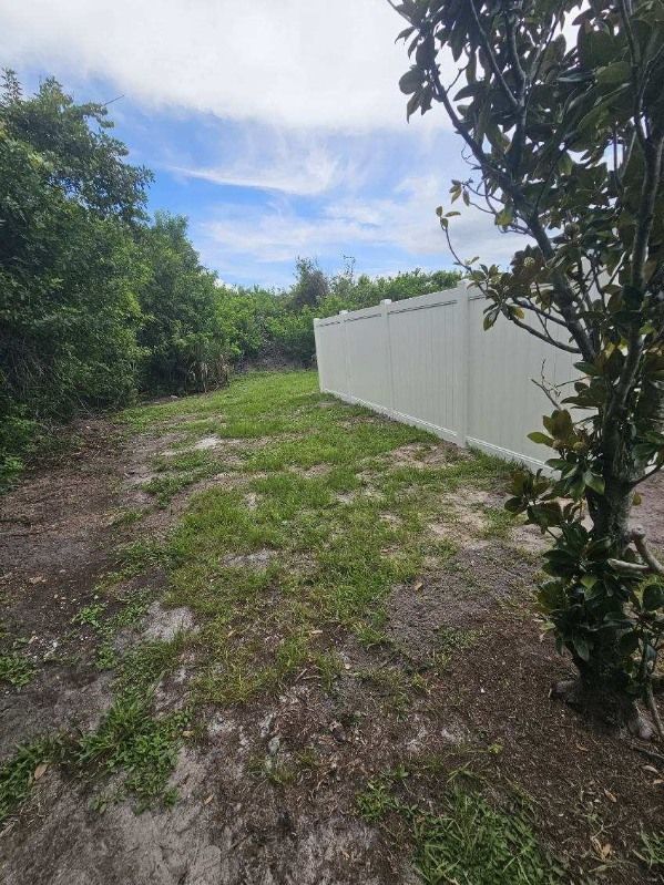Narrow grassy path beside a white fence, bordered by dense green trees under a partly cloudy sky