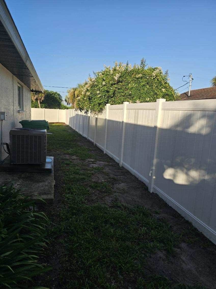 Narrow grassy side yard between a house and white fence, with stacked items near the wall.