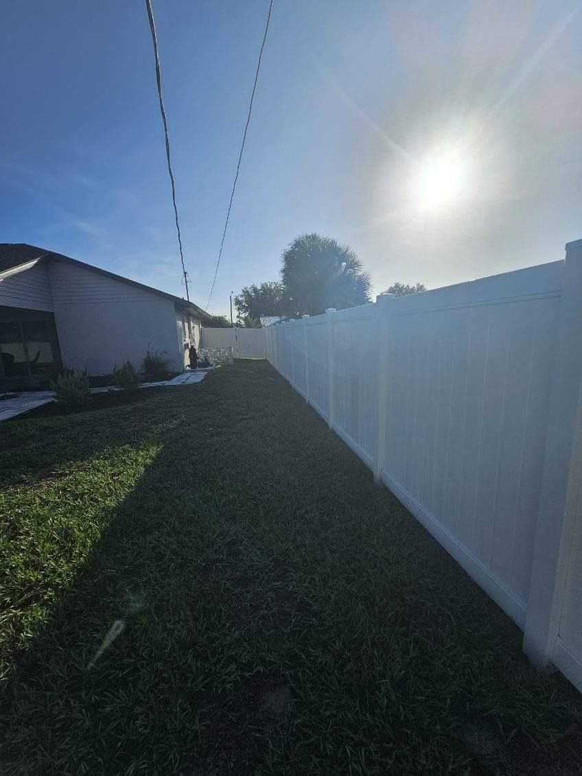 Narrow grassy alley beside a white fence and house, with power lines overhead and bright sun ahead.