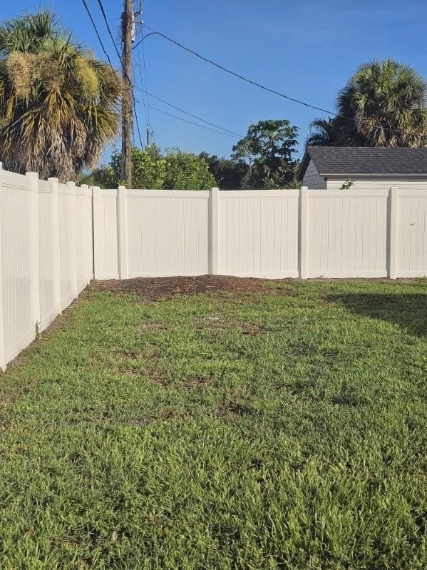 Small backyard with green grass, bare soil patch, and white privacy fence under a blue sky.