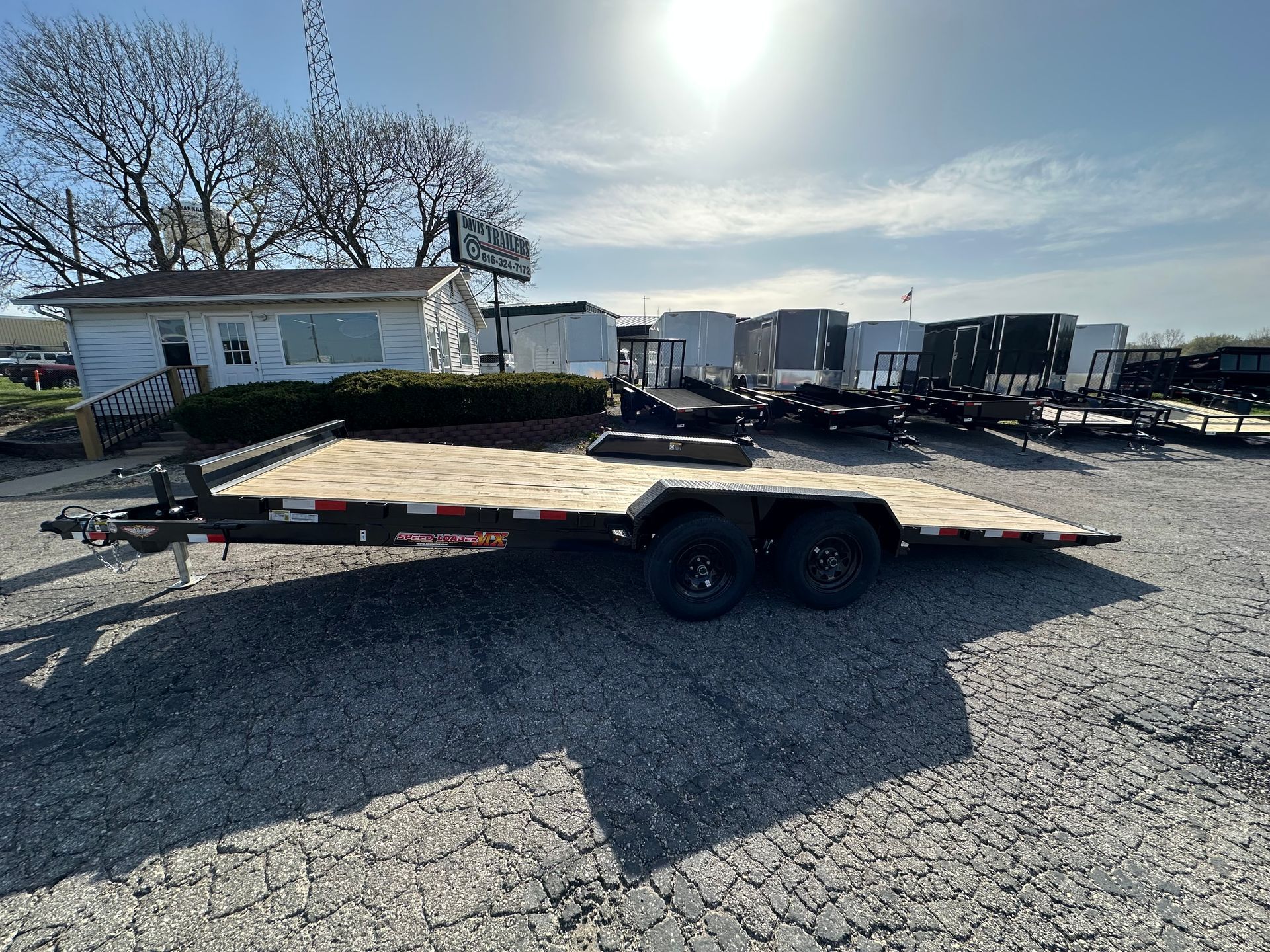 A flatbed trailer with a wooden deck is parked next to a white trailer.