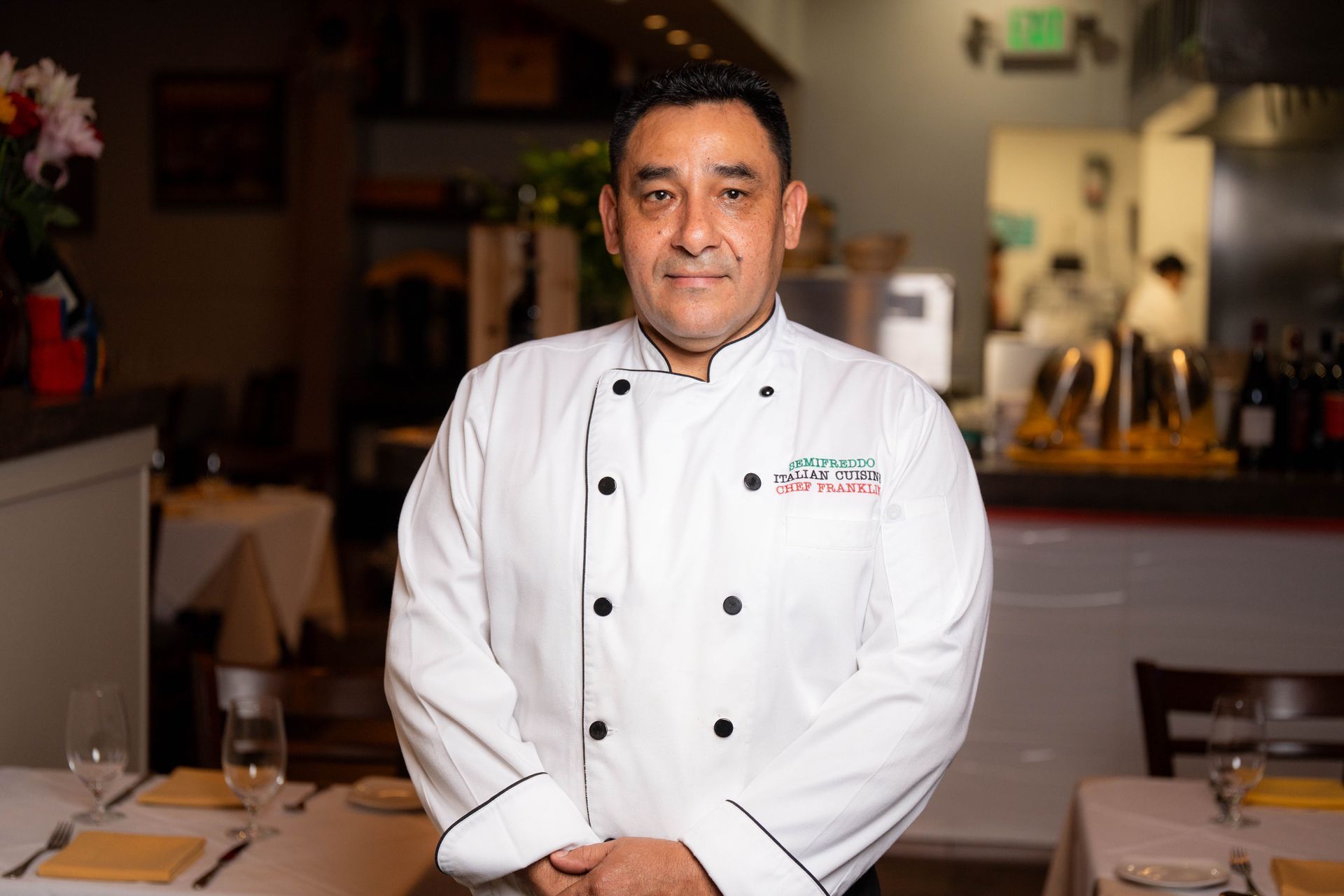 Chef standing in a restaurant, wearing a white coat, smiling. The background includes tables, flowers, and kitchen staff.