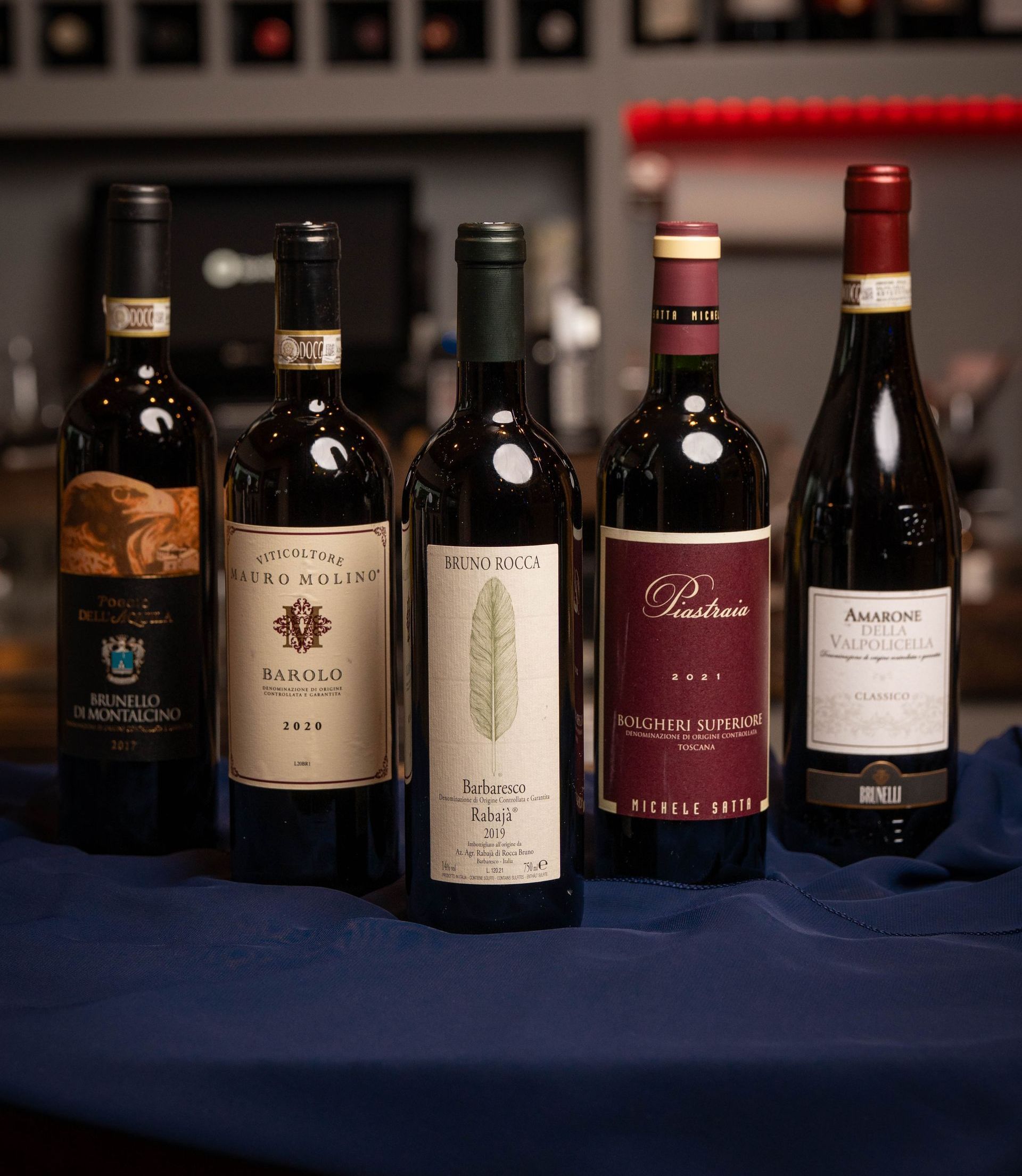 Six dark bottles of wine with colorful, patterned labels and bottle caps, arranged on a patterned tablecloth.