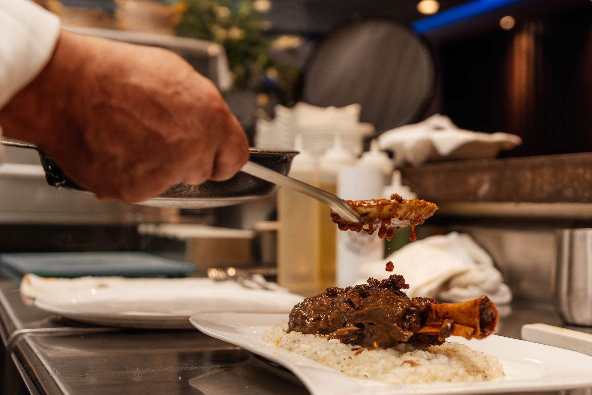 Chef plating a braised meat dish with rice on a white plate in a kitchen, pouring sauce.