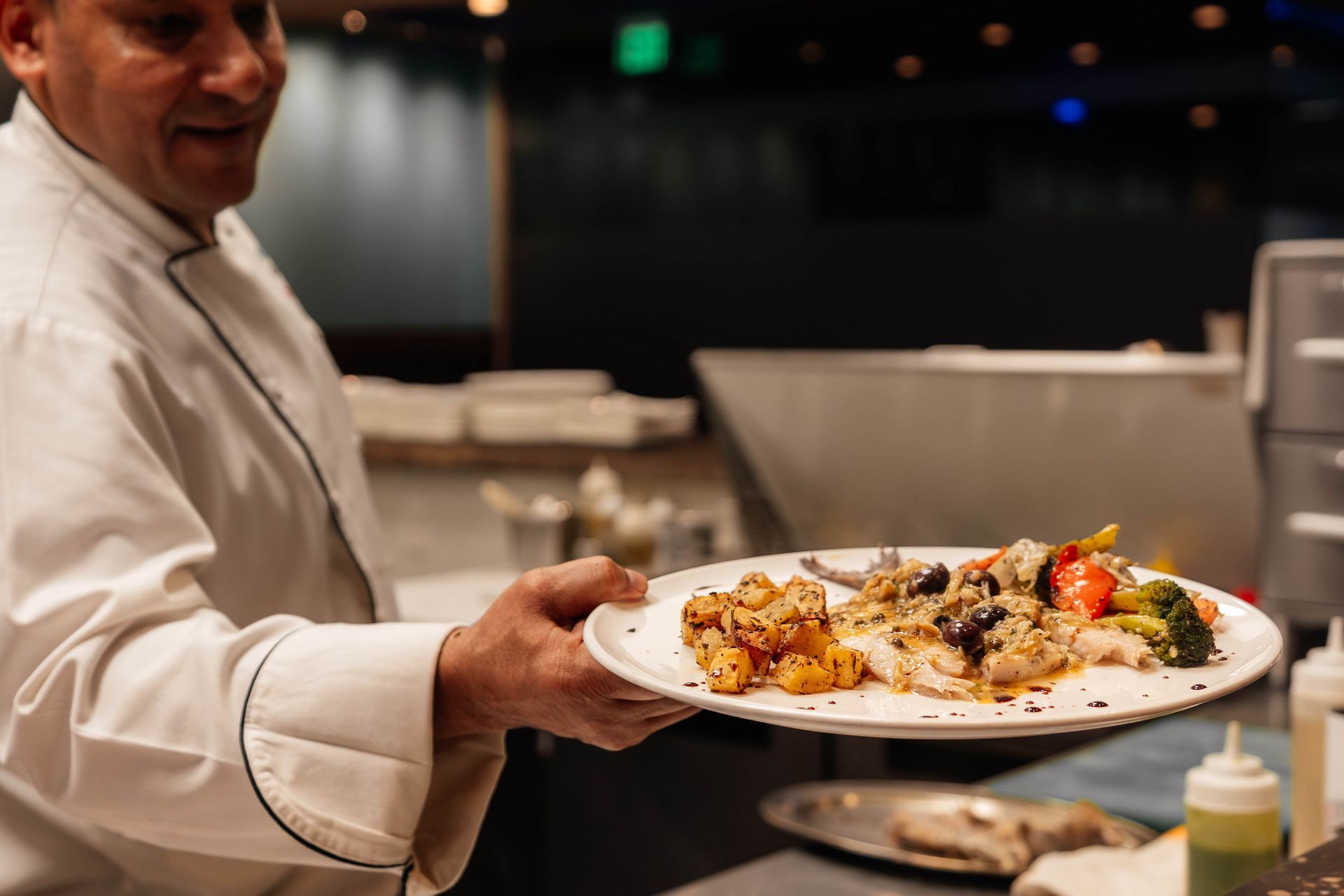 Chef holding a plate of food in a kitchen. Food includes potatoes, vegetables, and chicken.