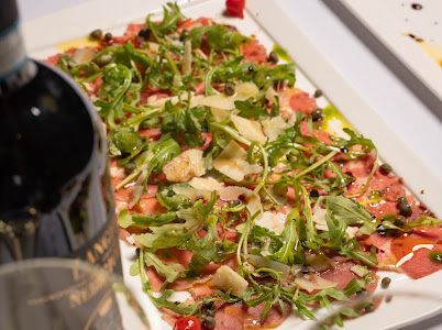 Carpaccio appetizer on a white platter, topped with arugula, parmesan, and capers, with a wine bottle.