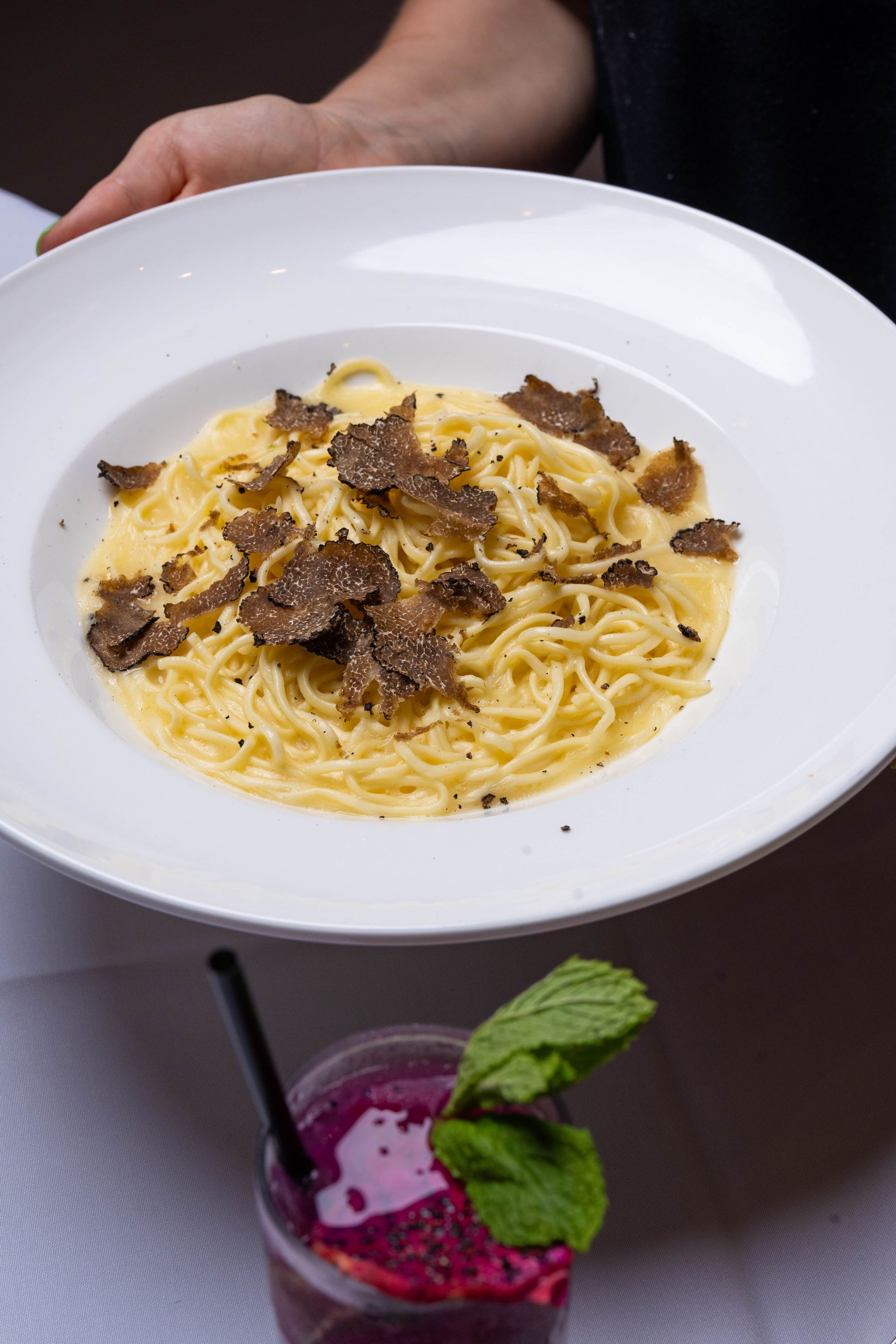Person holding a white plate of spaghetti with shaved black truffles, next to a cocktail.