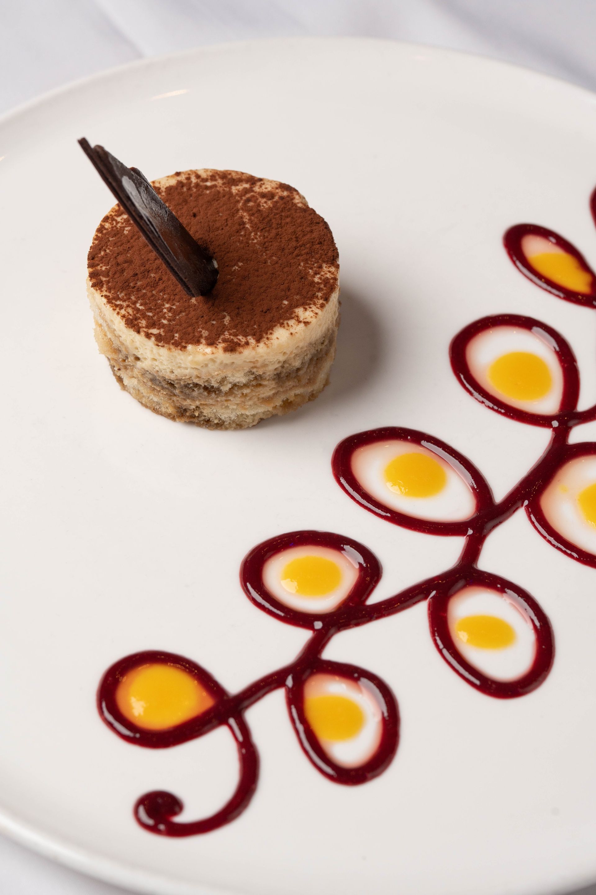Tiramisu dessert on a white plate, topped with cocoa and a chocolate stick, next to a decorative berry and mango sauce design.