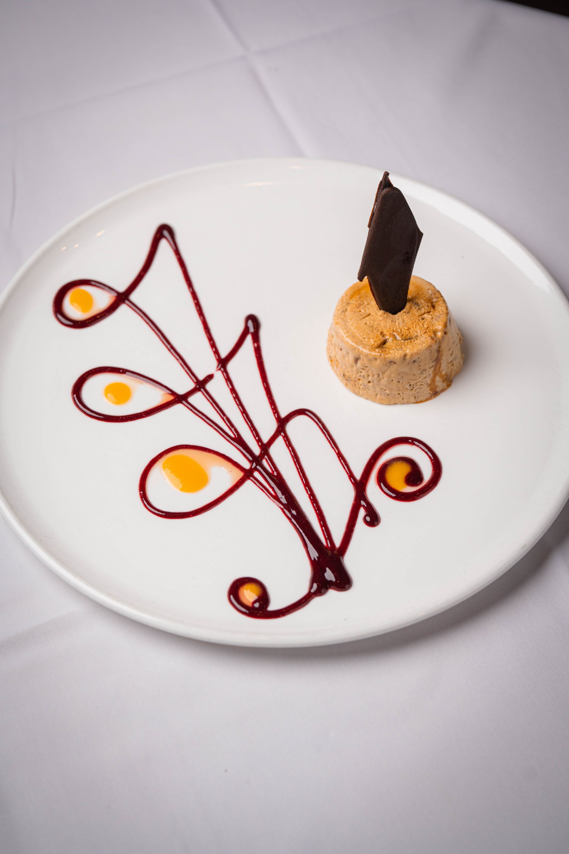 Dessert plate with a small circular mousse topped with a dark chocolate shard, and decorative sauce swirls in burgundy and orange.