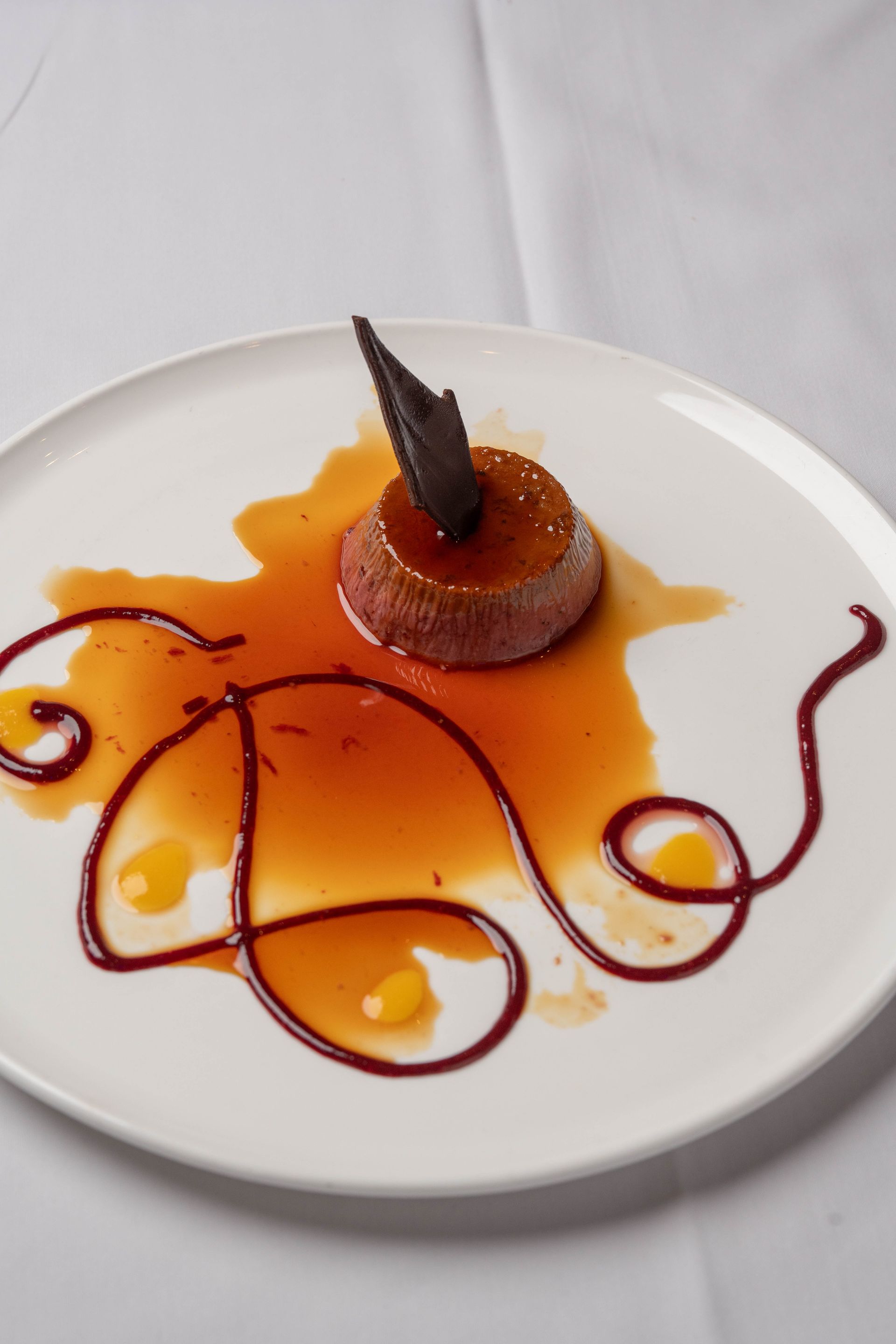 A dessert on a white plate with a caramel-glazed center, chocolate garnish, and decorative sauce.