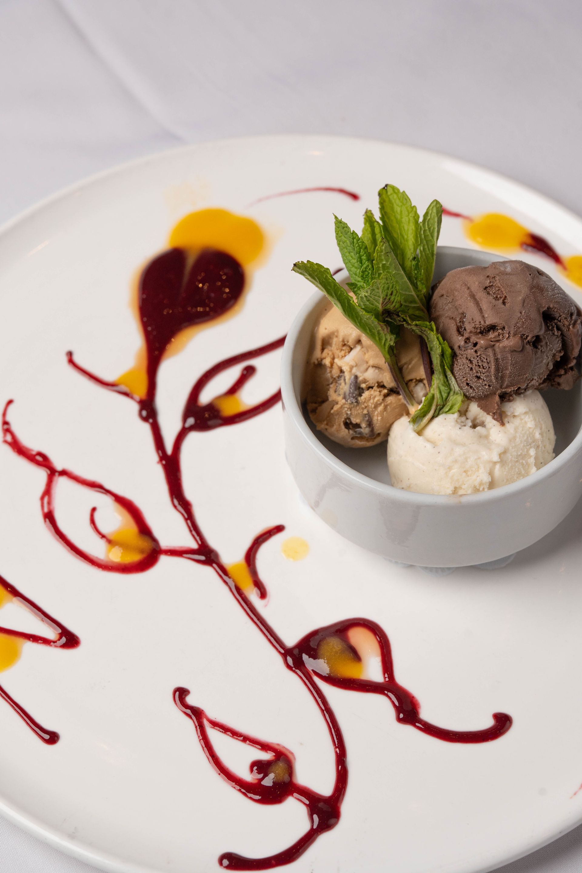 Three scoops of ice cream in a white bowl, decorated with a fruit puree design on a white plate. Mint sprigs adorn the ice cream.