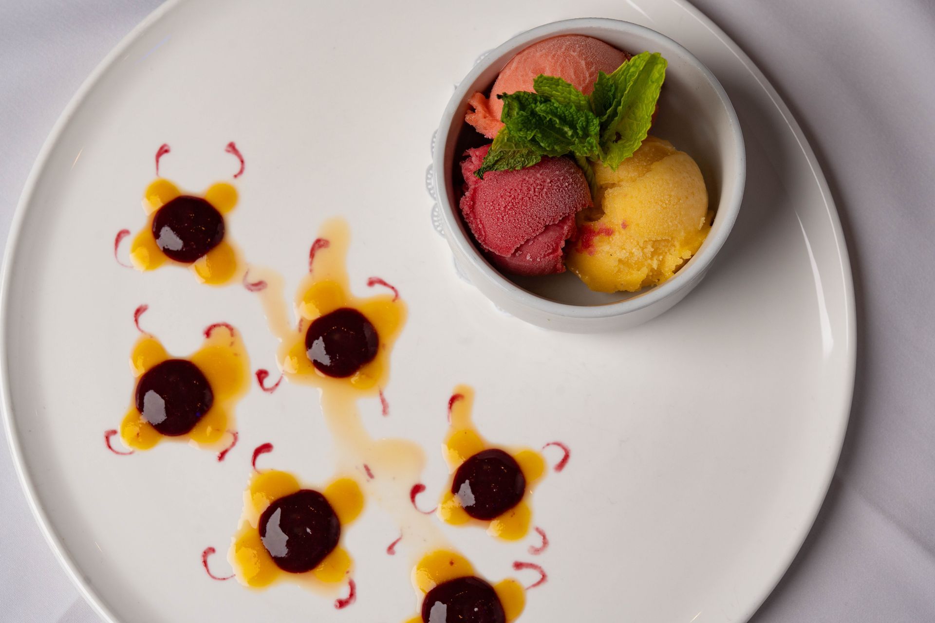 White plate with scoops of fruit sorbet in a small bowl garnished with mint. Decorative sauce art and berry dots on the plate.