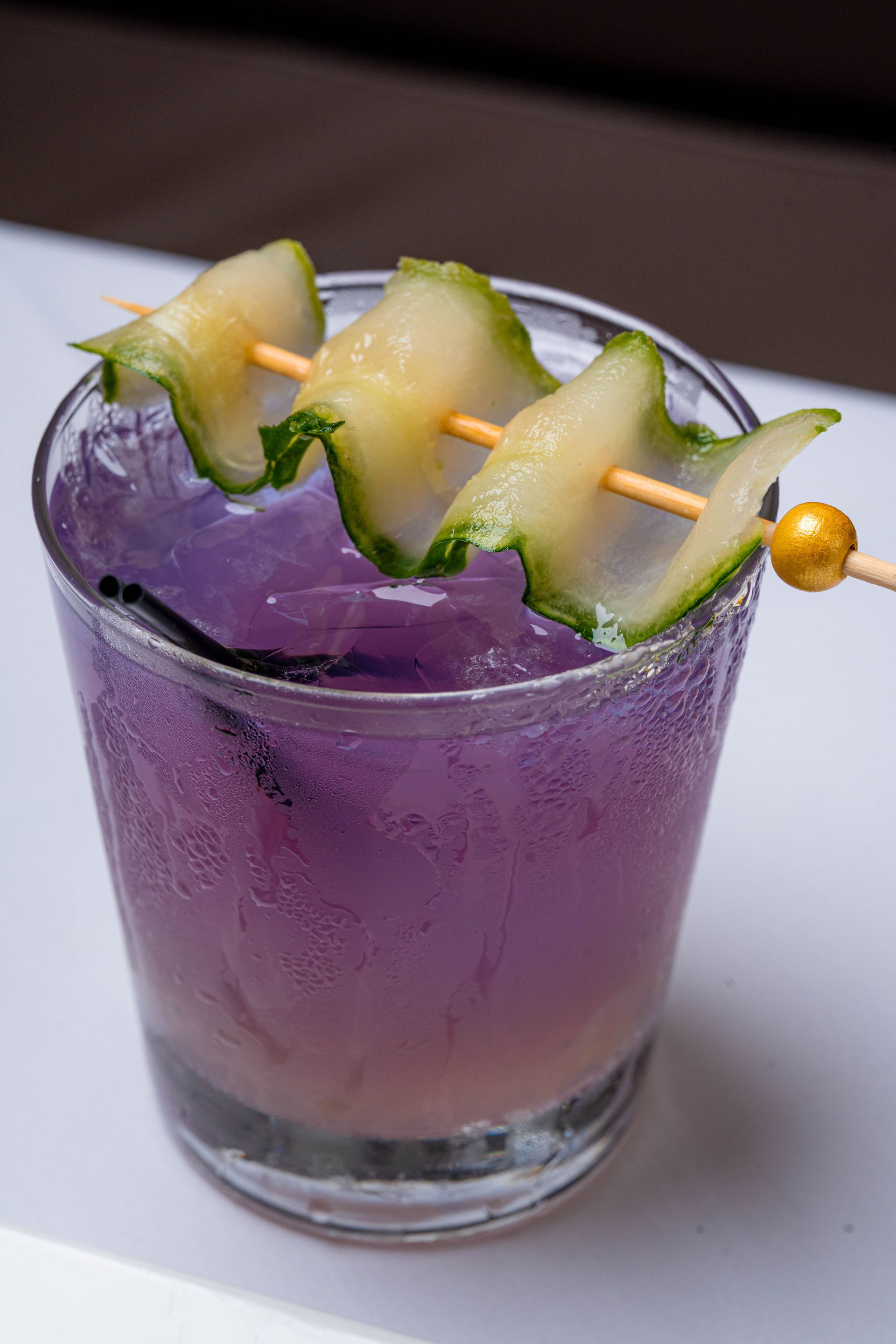 Purple cocktail in a glass, garnished with cucumber slices on a skewer. The drink sits on a white surface.