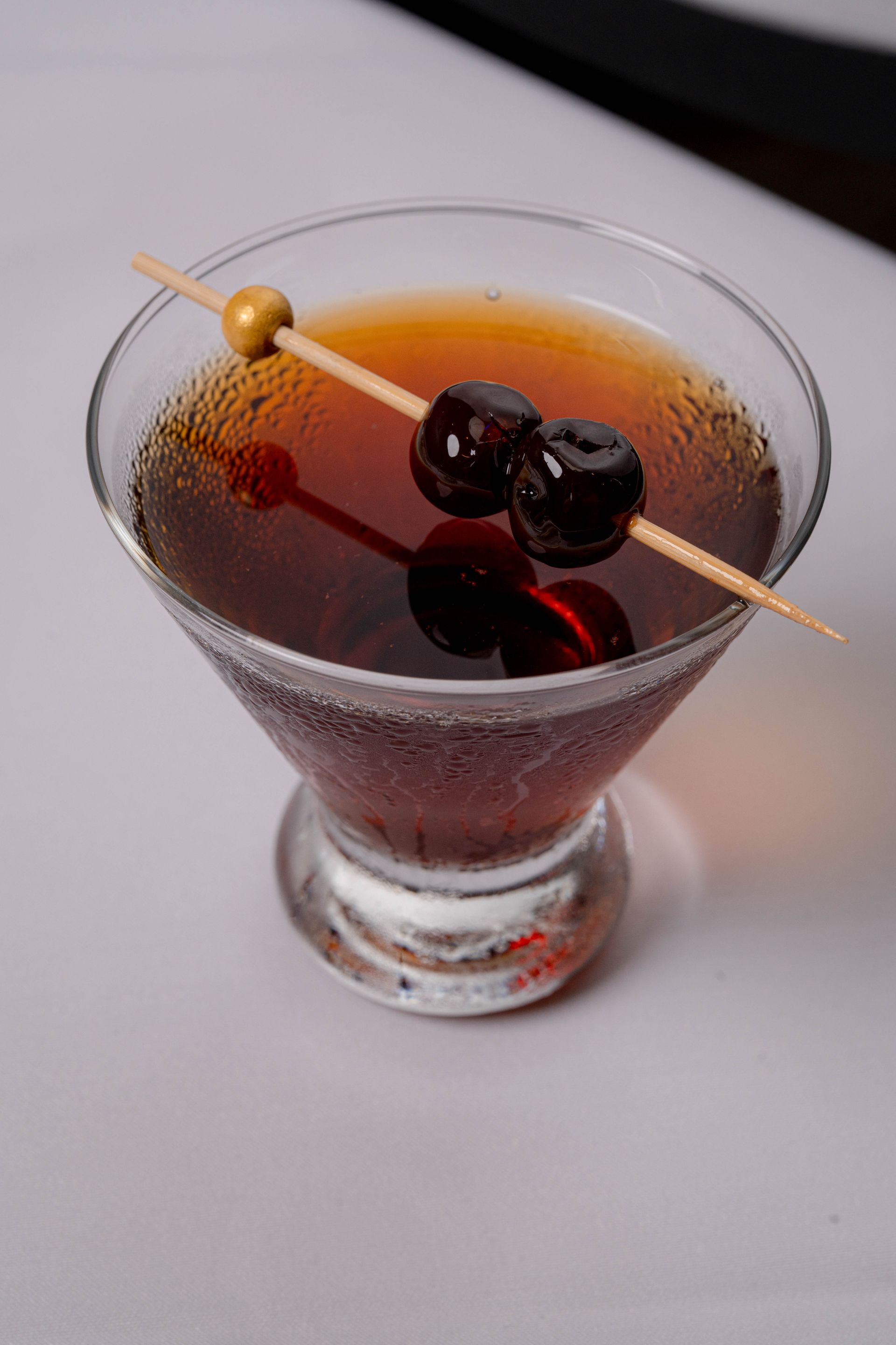 A dark brown cocktail in a martini glass, garnished with cherries on a skewer.