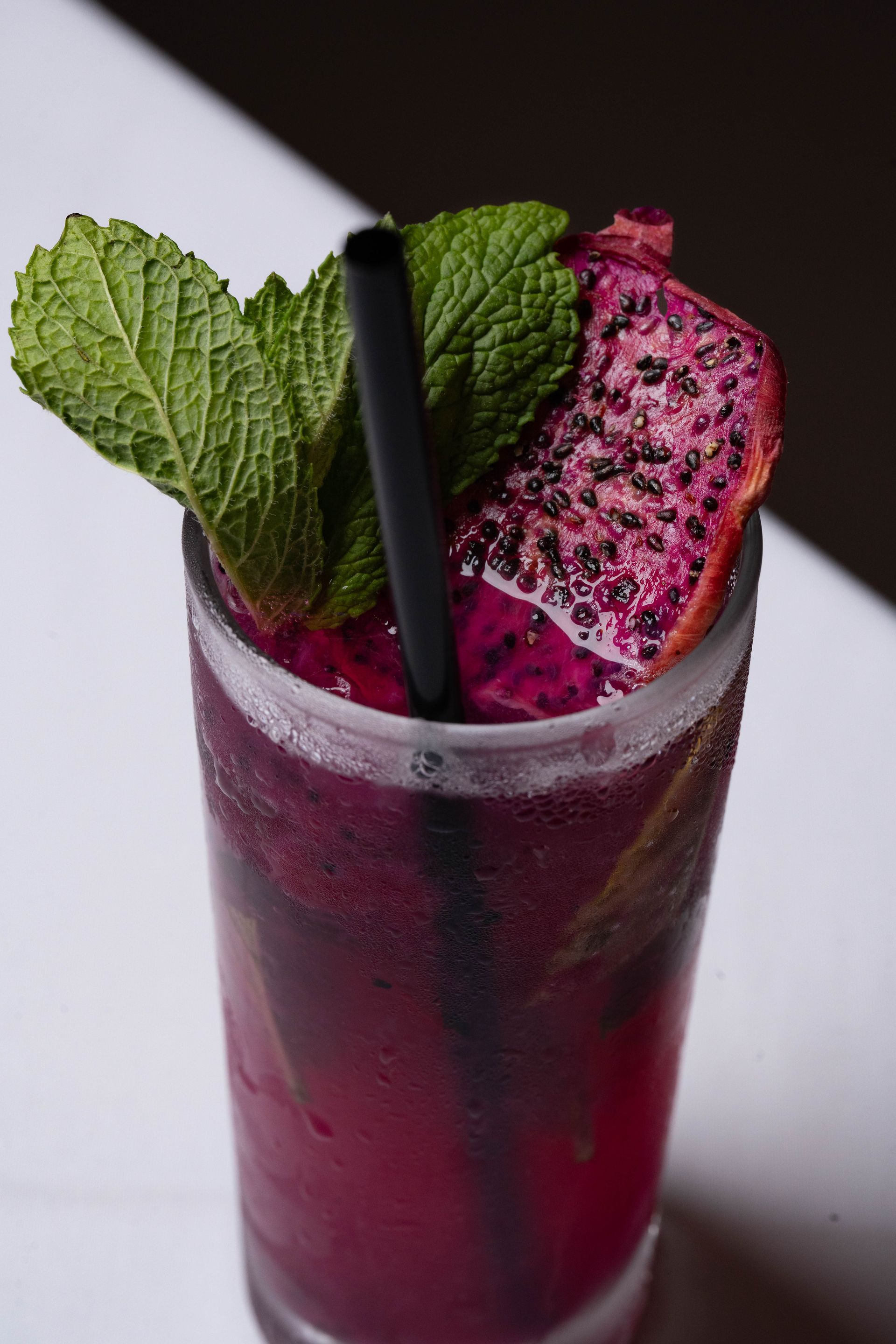 A vibrant pink cocktail in a tall glass, garnished with mint and a dragon fruit slice, with a black straw.