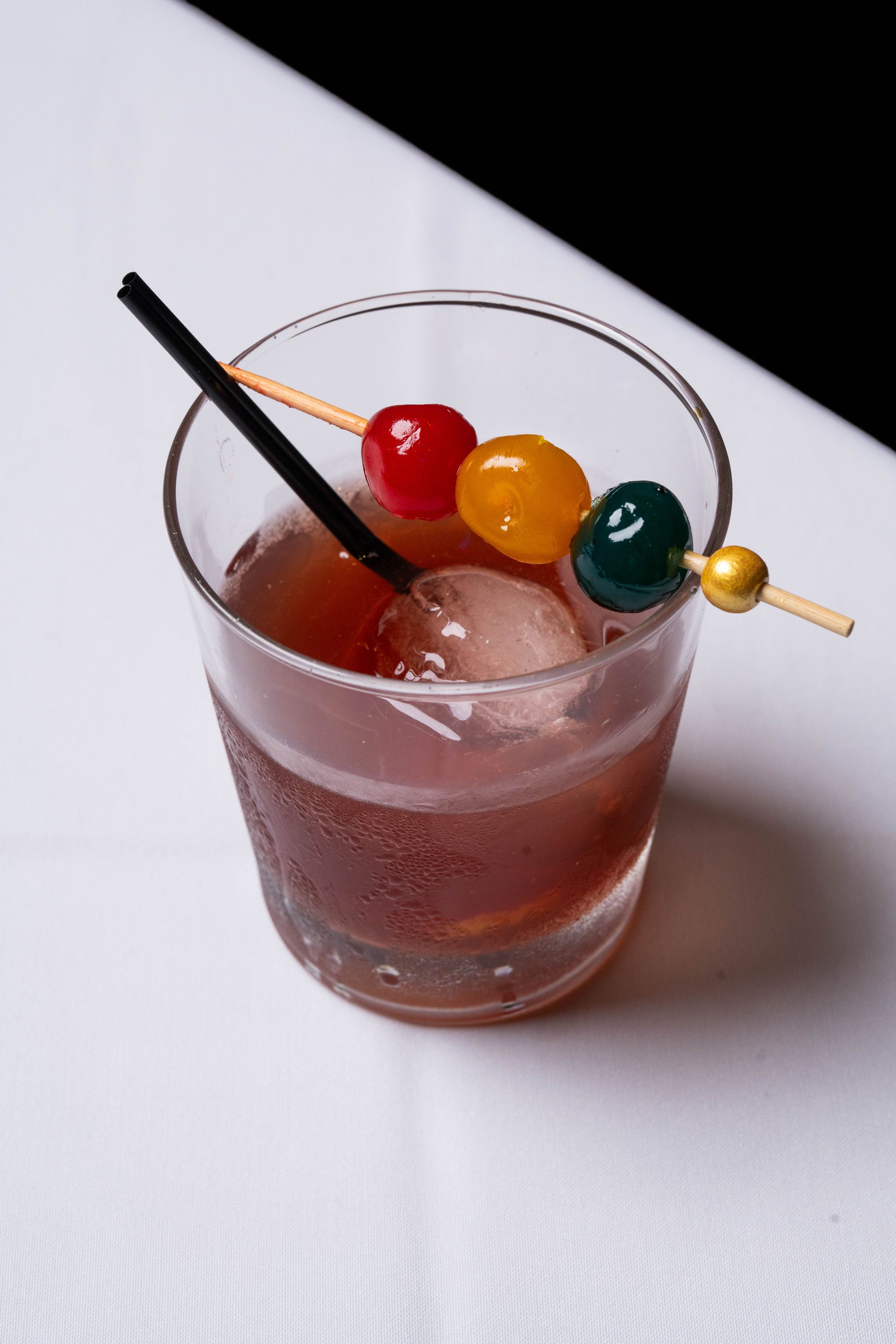 A cocktail in a rocks glass, garnished with cherries on a skewer and a straw, on a white surface with a black backdrop.