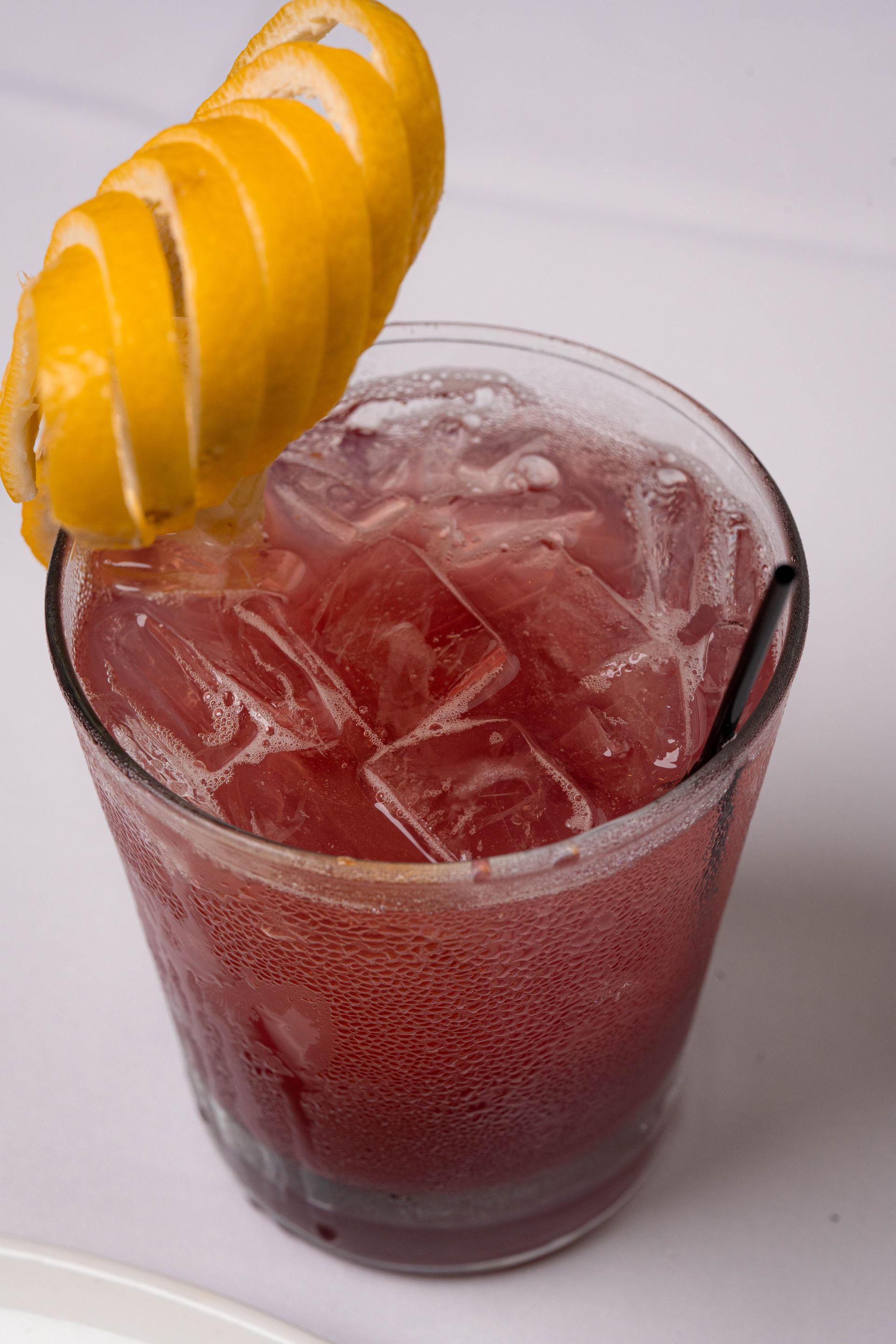 Red cocktail in a glass with ice, garnished with a lemon peel.