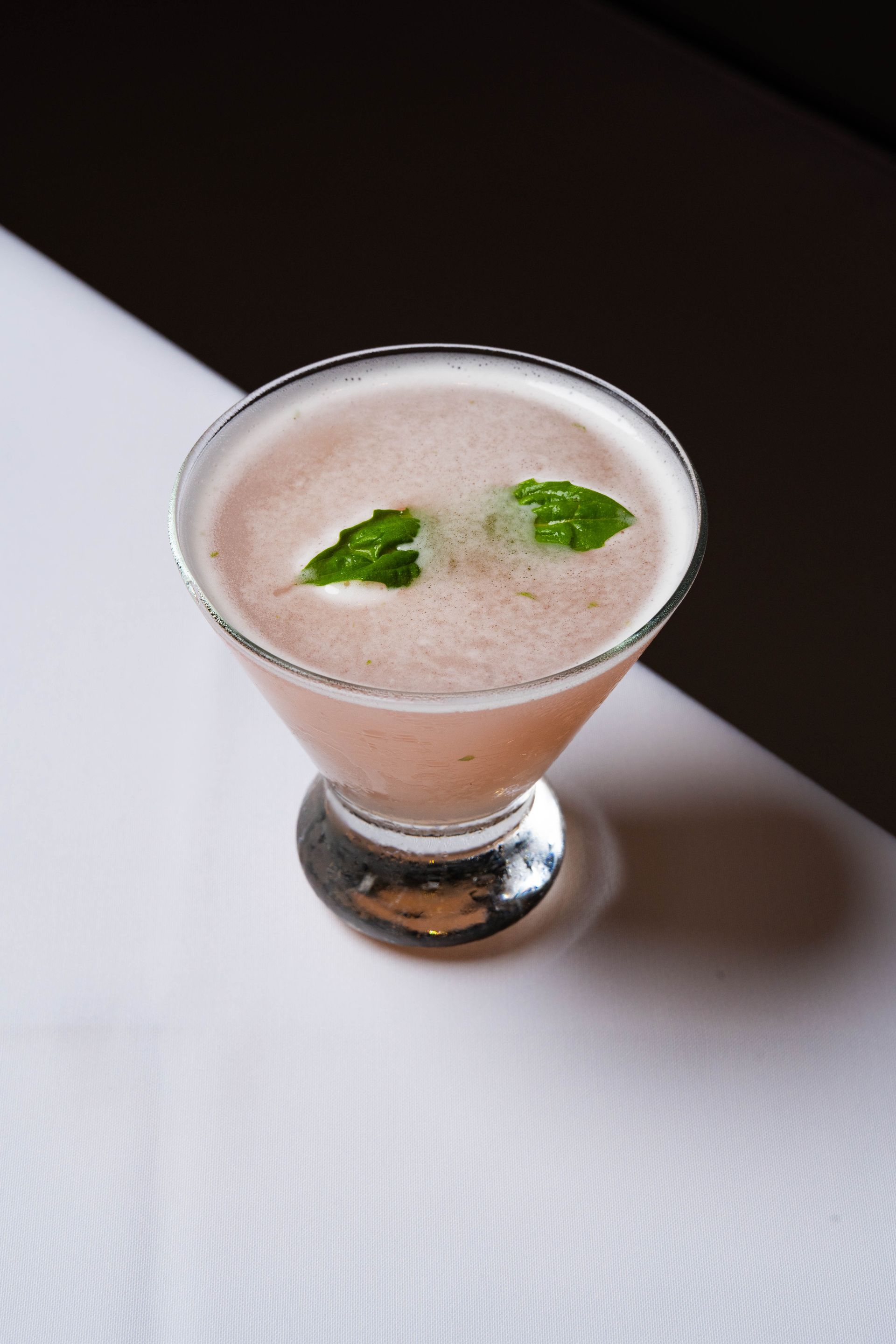 Pink cocktail in a stemmed glass, garnished with mint leaves, on a black and white surface.