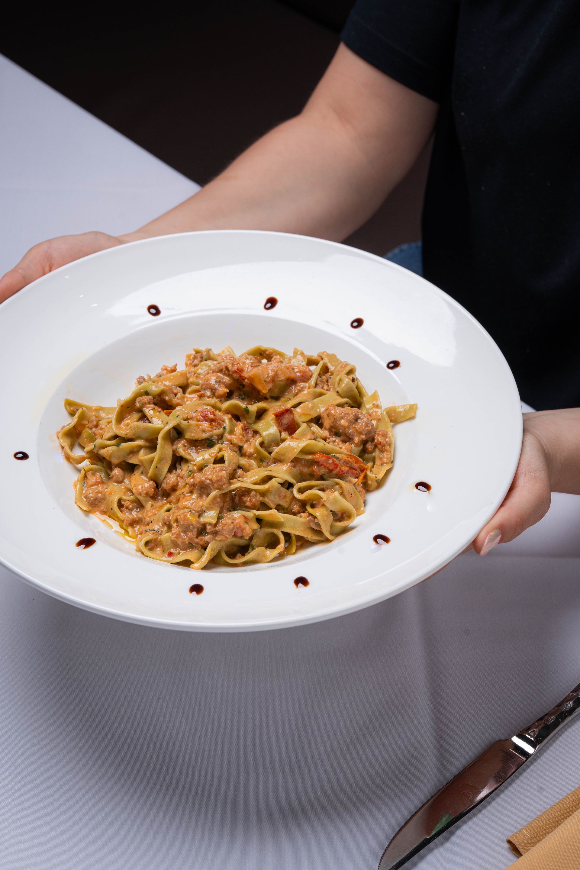 Person holding a white plate of pasta with a tomato-based sauce, garnished with balsamic vinegar drops.