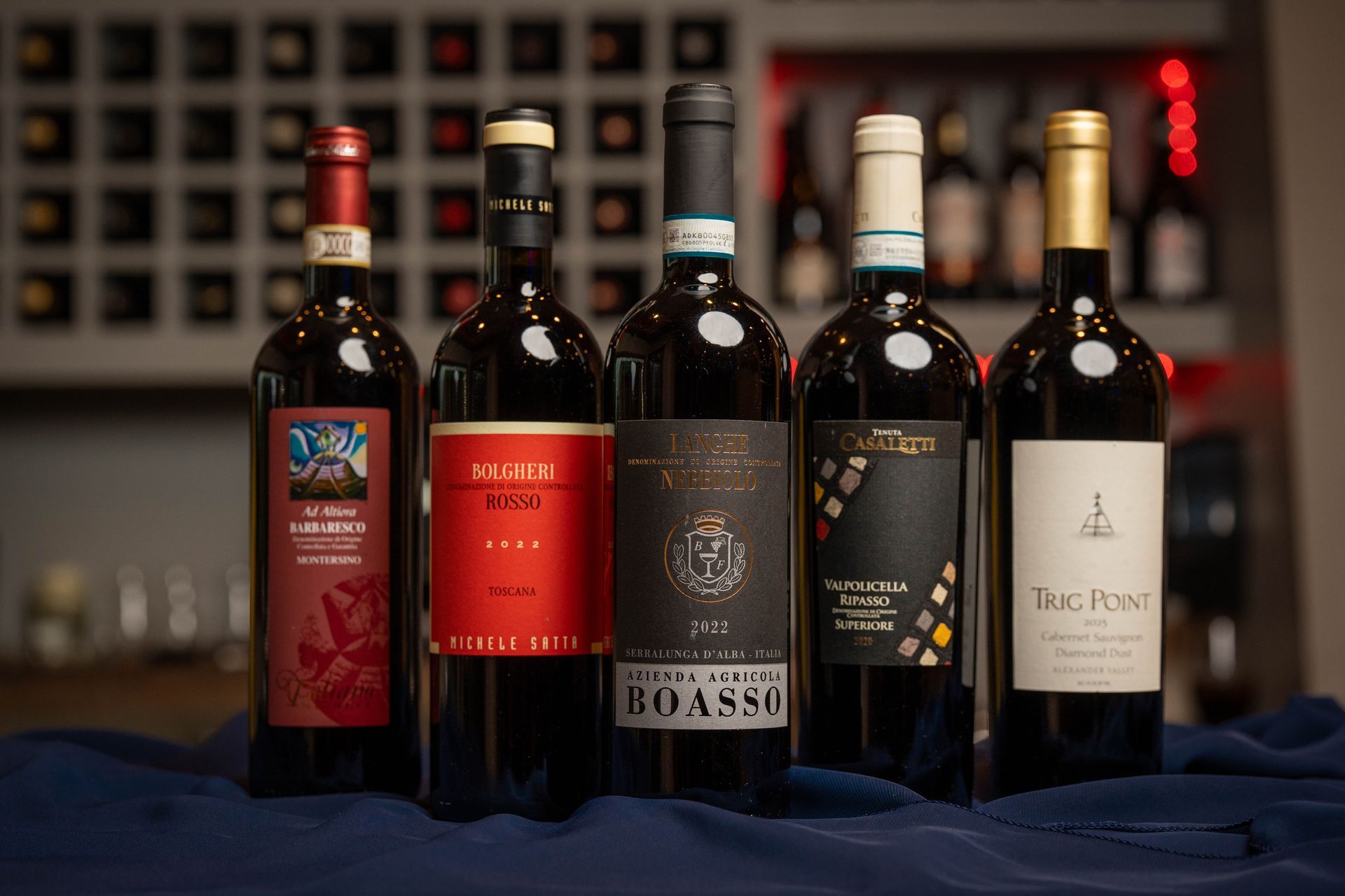 Five bottles of red wine on a blue cloth-covered surface. Labels vary, with colors including red, black, and gold.
