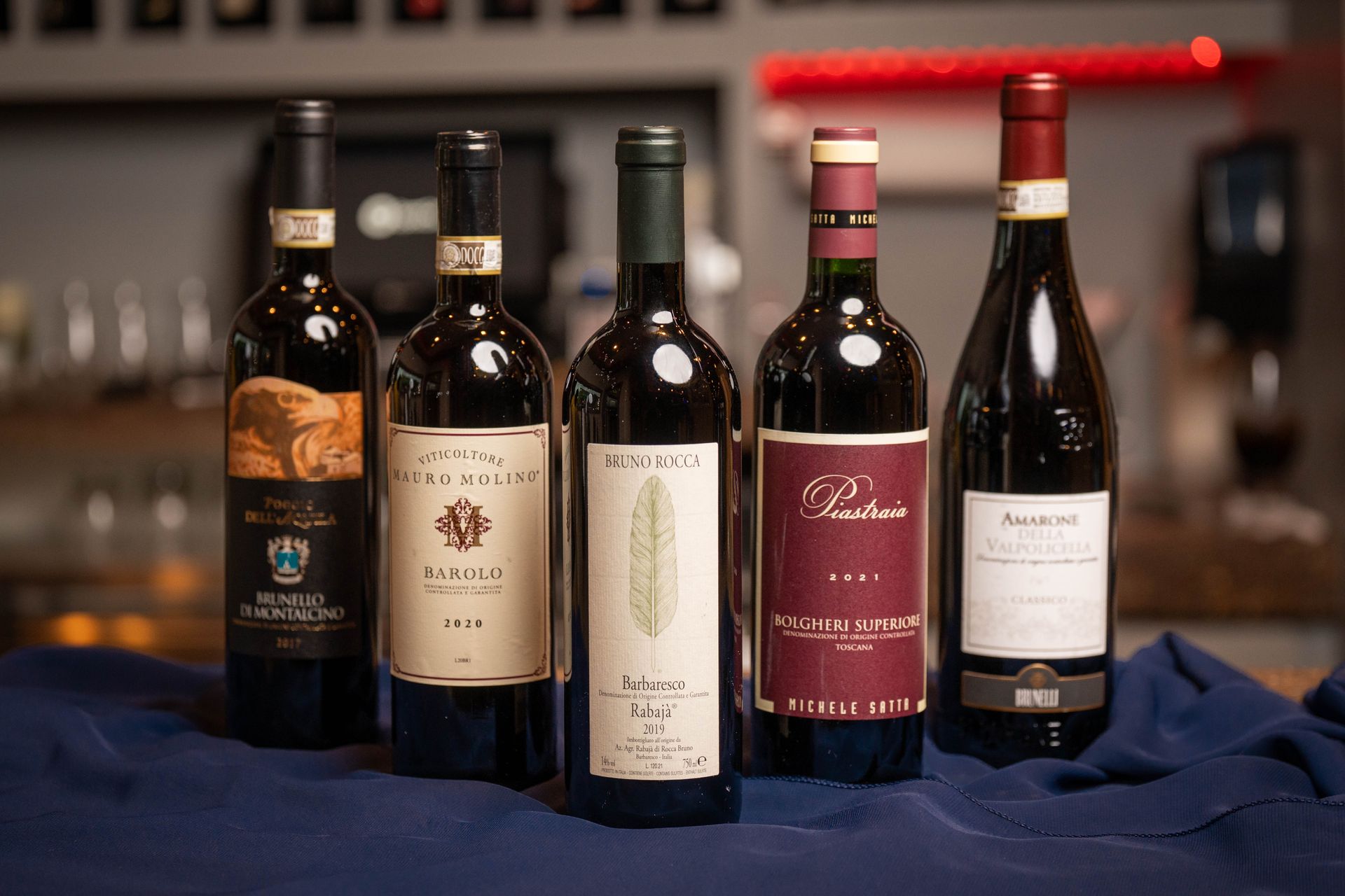 Five bottles of red wine stand on a blue cloth in a dimly lit setting. Labels vary, featuring text and designs.