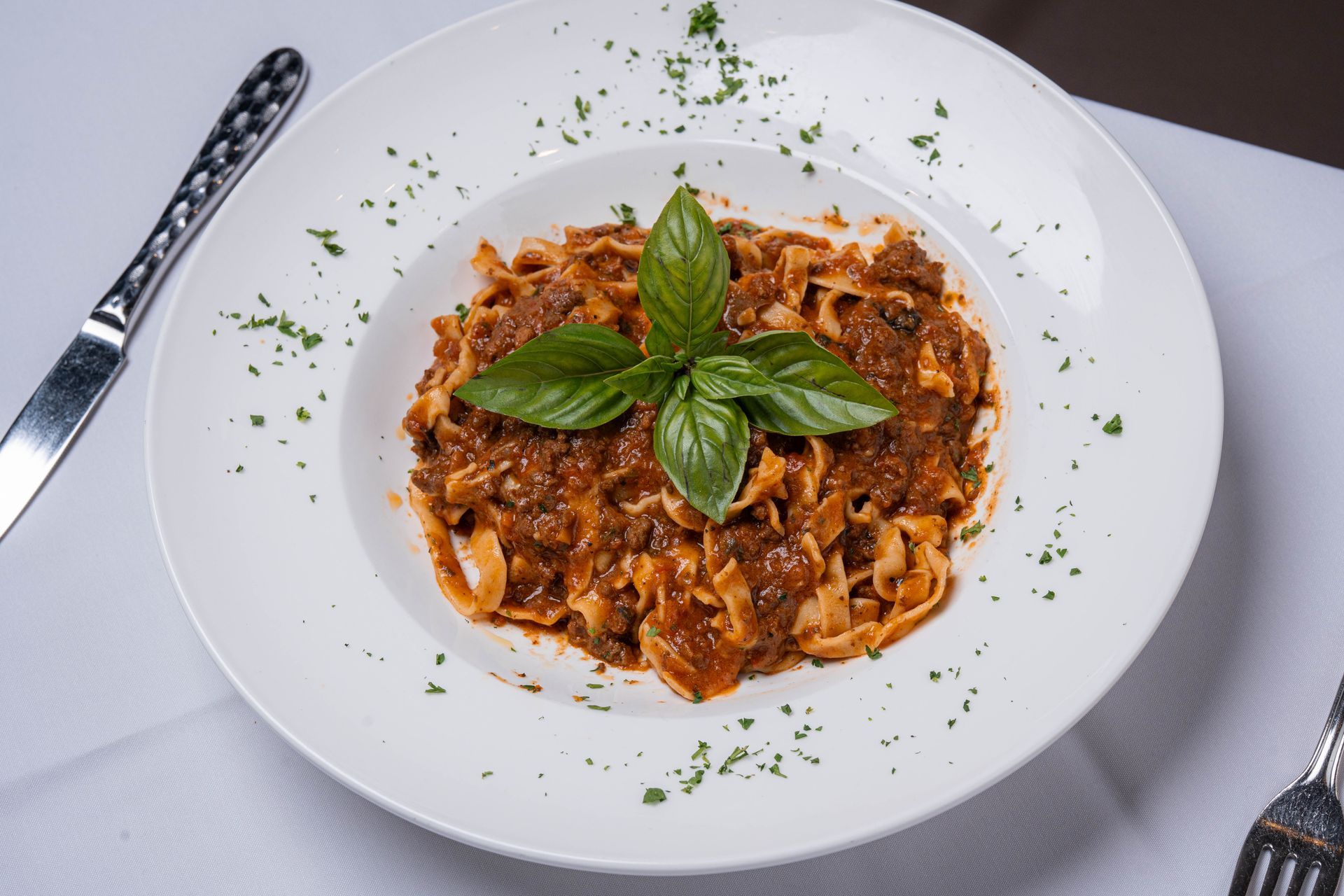 Fettuccine pasta with meat sauce, garnished with basil leaves and parsley, served on a white plate with silverware.