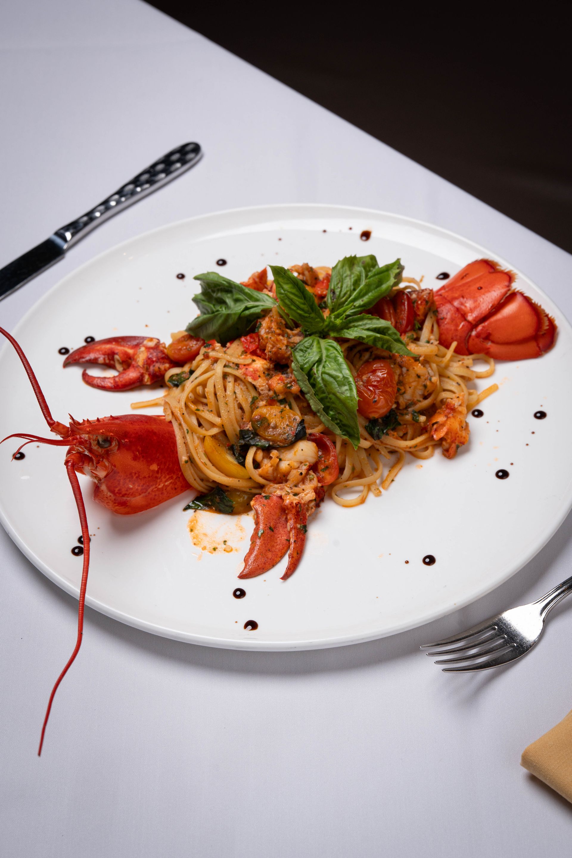 Lobster pasta dish with a whole lobster, basil, and balsamic glaze on a white plate on a white tablecloth.