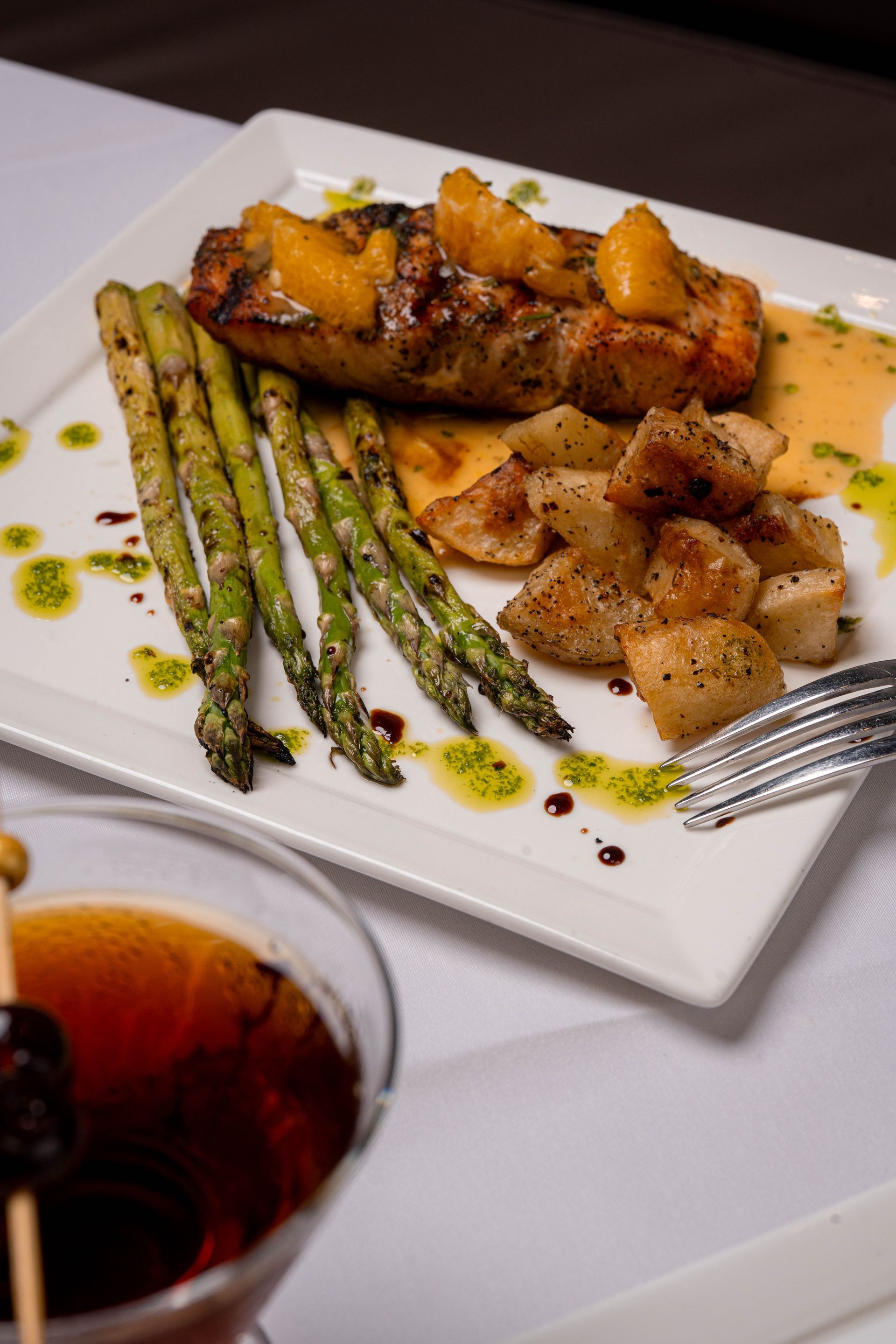 Grilled salmon with orange topping, asparagus, and potatoes on a white plate, with a cocktail in the foreground.